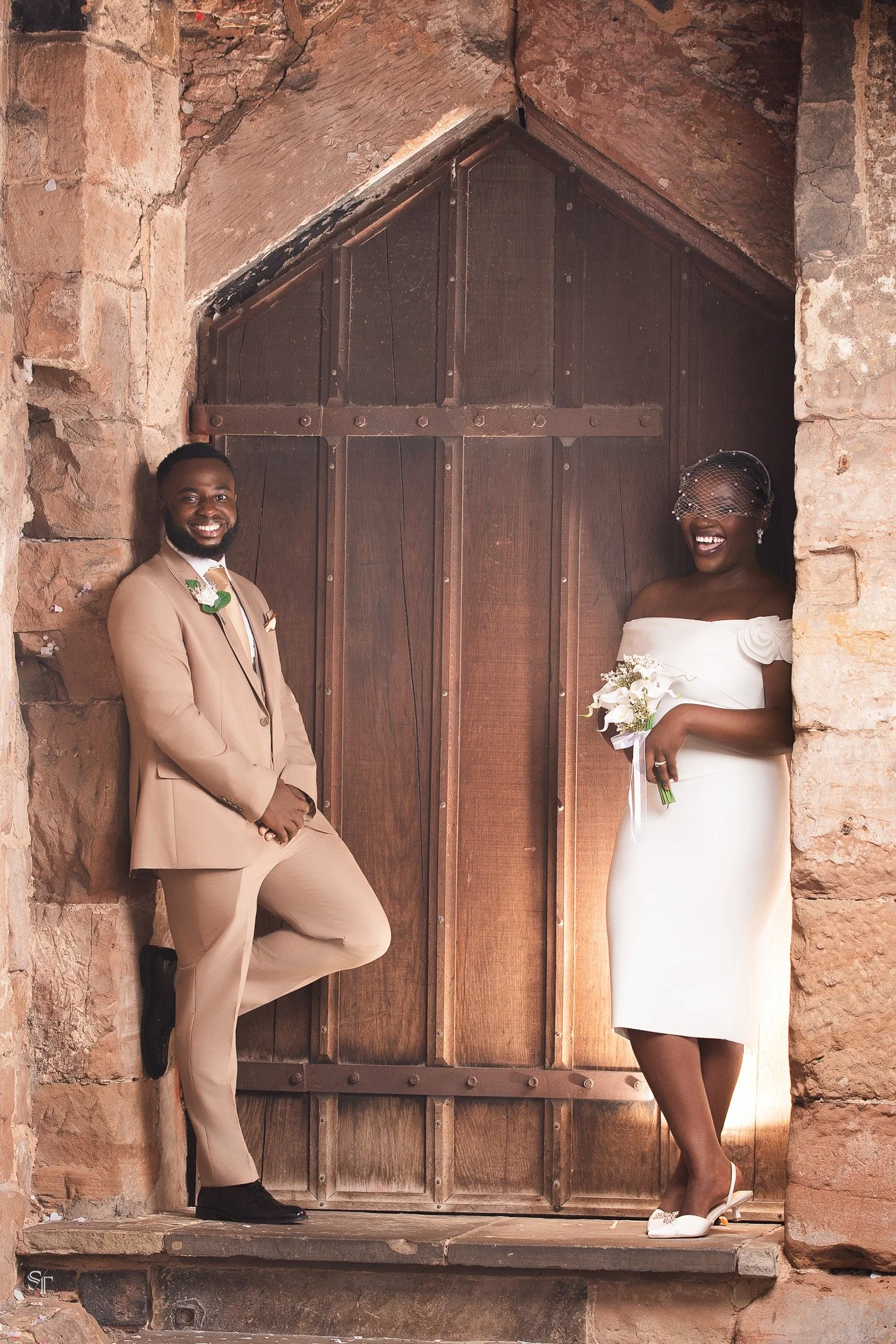 A smiling man in a beige suit and black shoes leaning against a stone wall, and a smiling woman in a white dress holding a bouquet, standing in front of a large wooden door set in an arched stone entrance.
