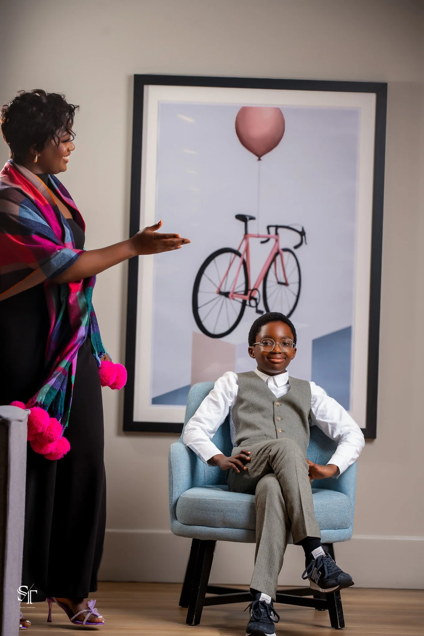 A young boy in a gray vest and pants sitting in a light blue armchair, smiling, with a woman standing nearby gesturing towards him, indoors with a large framed artwork of a pink bicycle and a balloon in the background.