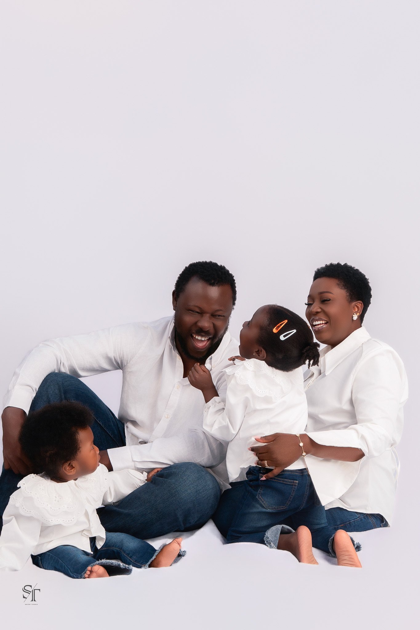 Family of four, two adults and two children, sitting and standing together on a white background, wearing white shirts, smiling and enjoying each other's company.
