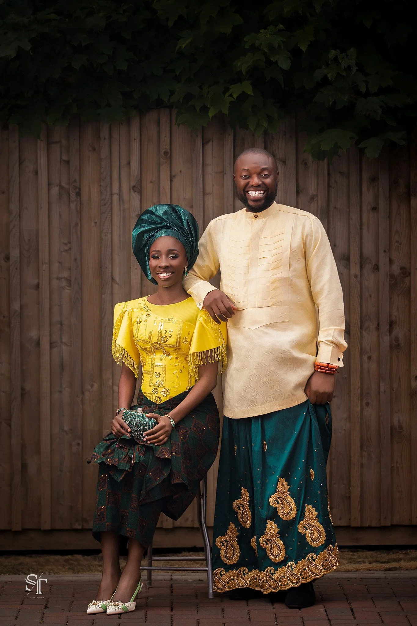 A man and woman dressed in traditional African attire standing outdoors in front of a wooden fence and leafy green tree. The woman is wearing a yellow dress, a green headwrap, and holding a clutch, while the man is wearing a cream shirt and a pattern