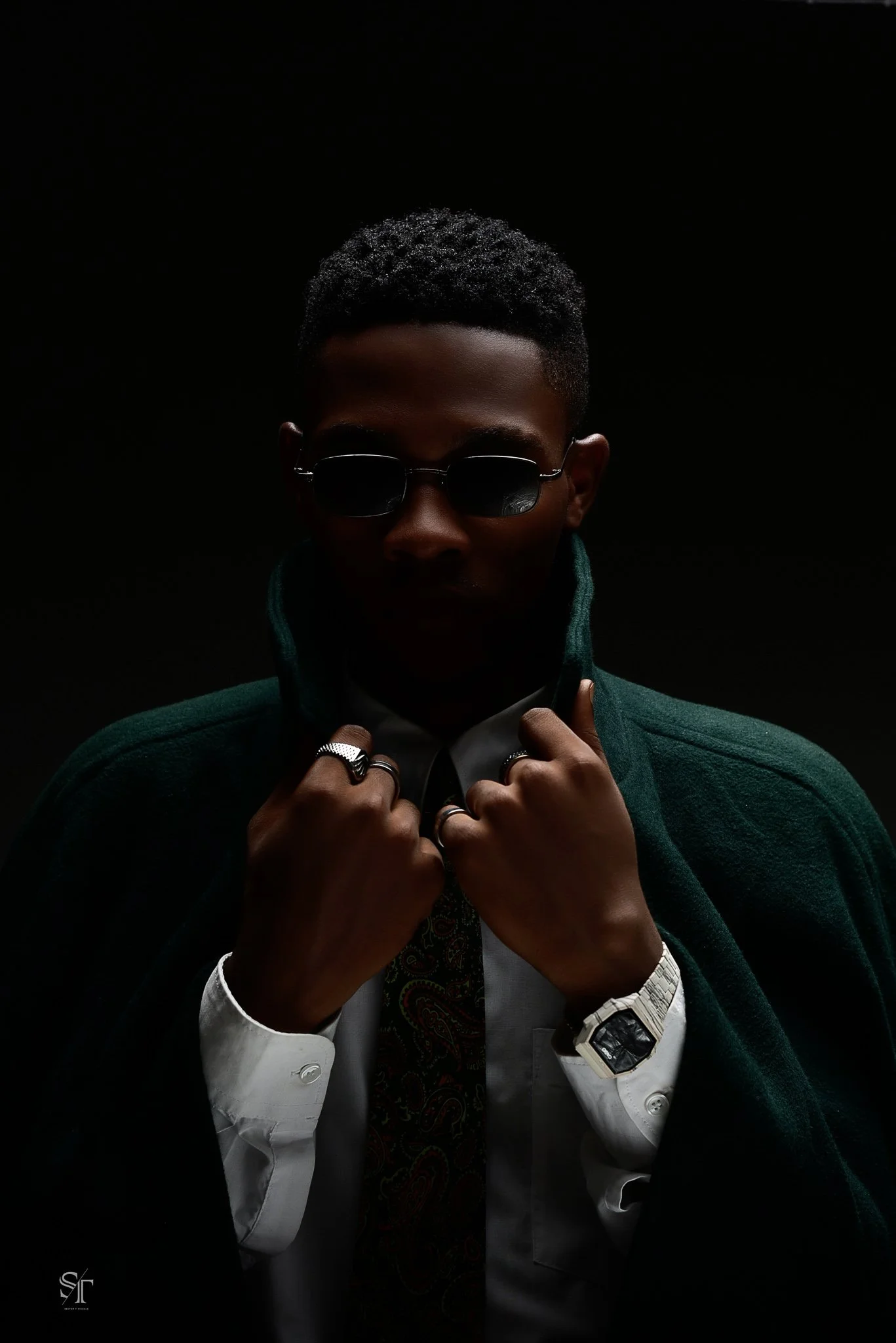 A man wearing sunglasses, a dark green jacket, a white shirt, rings, a watch, and a patterned tie, posing against a black background with dramatic lighting.