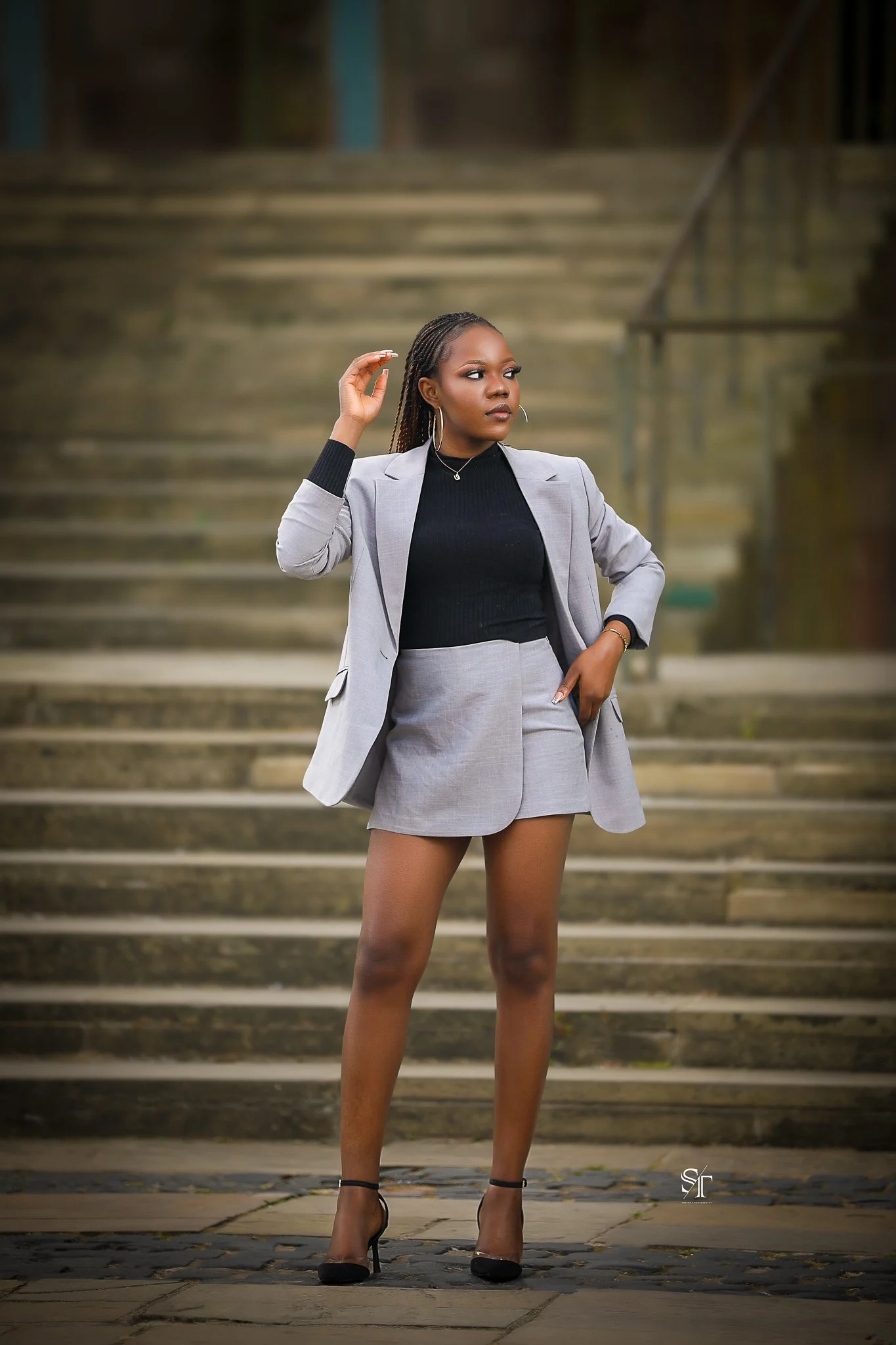 A stylish woman in a gray blazer and shorts, black top, black high heels, standing outdoors on stone pavement with stairs behind her, black earrings, and hair in braids.