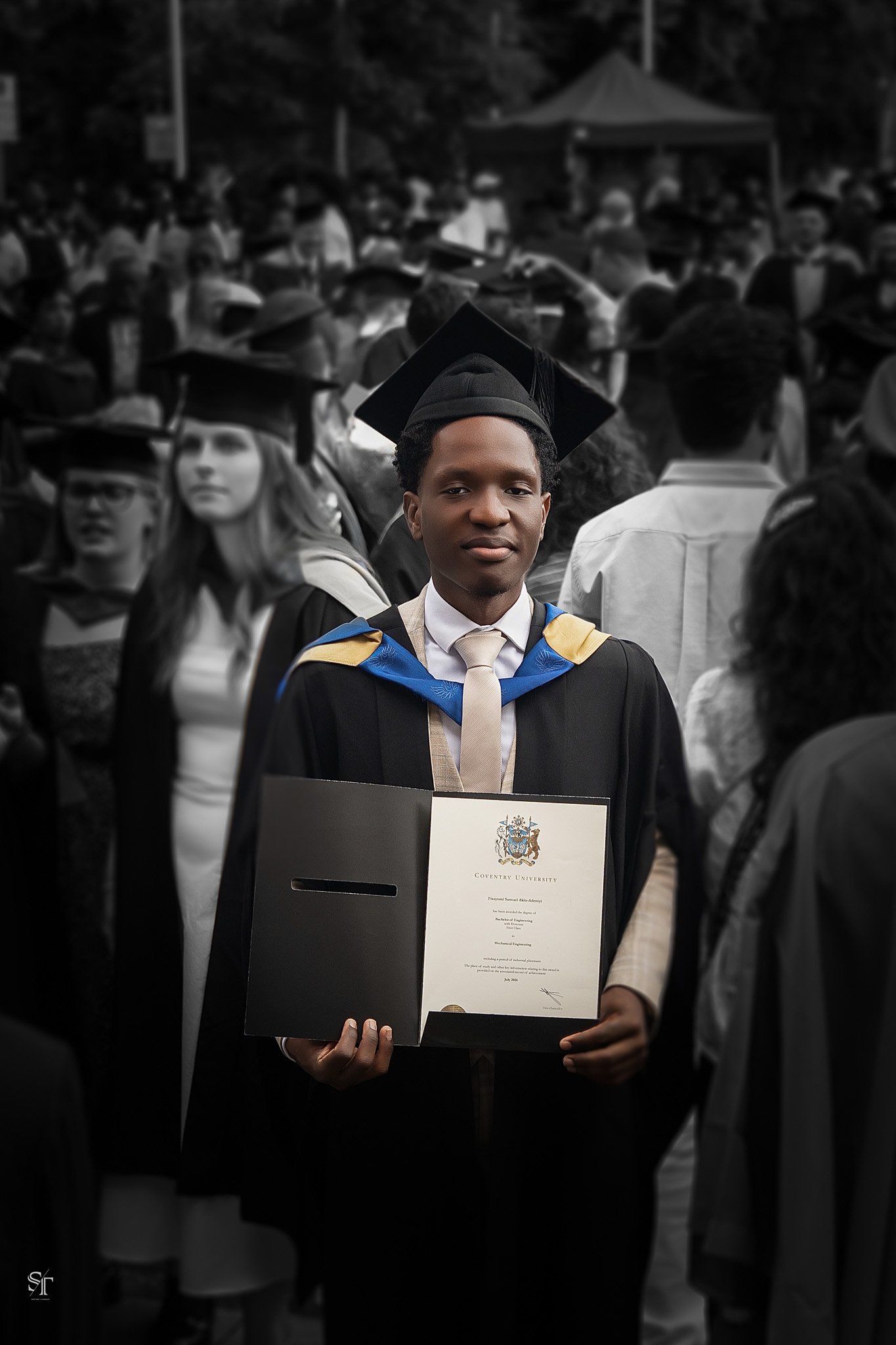A young man in a graduation cap and gown holding a diploma at graduation ceremony. The background shows a crowd of graduates, mostly in black and white, with some in color, wearing caps and gowns.
