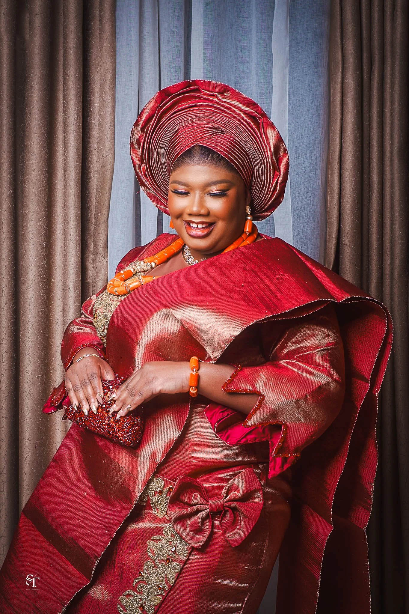 A woman dressed in traditional Nigerian red and gold attire with a matching gele (headwrap), earrings, necklace, bracelet, and holding a clutch purse, smiling in front of curtains.