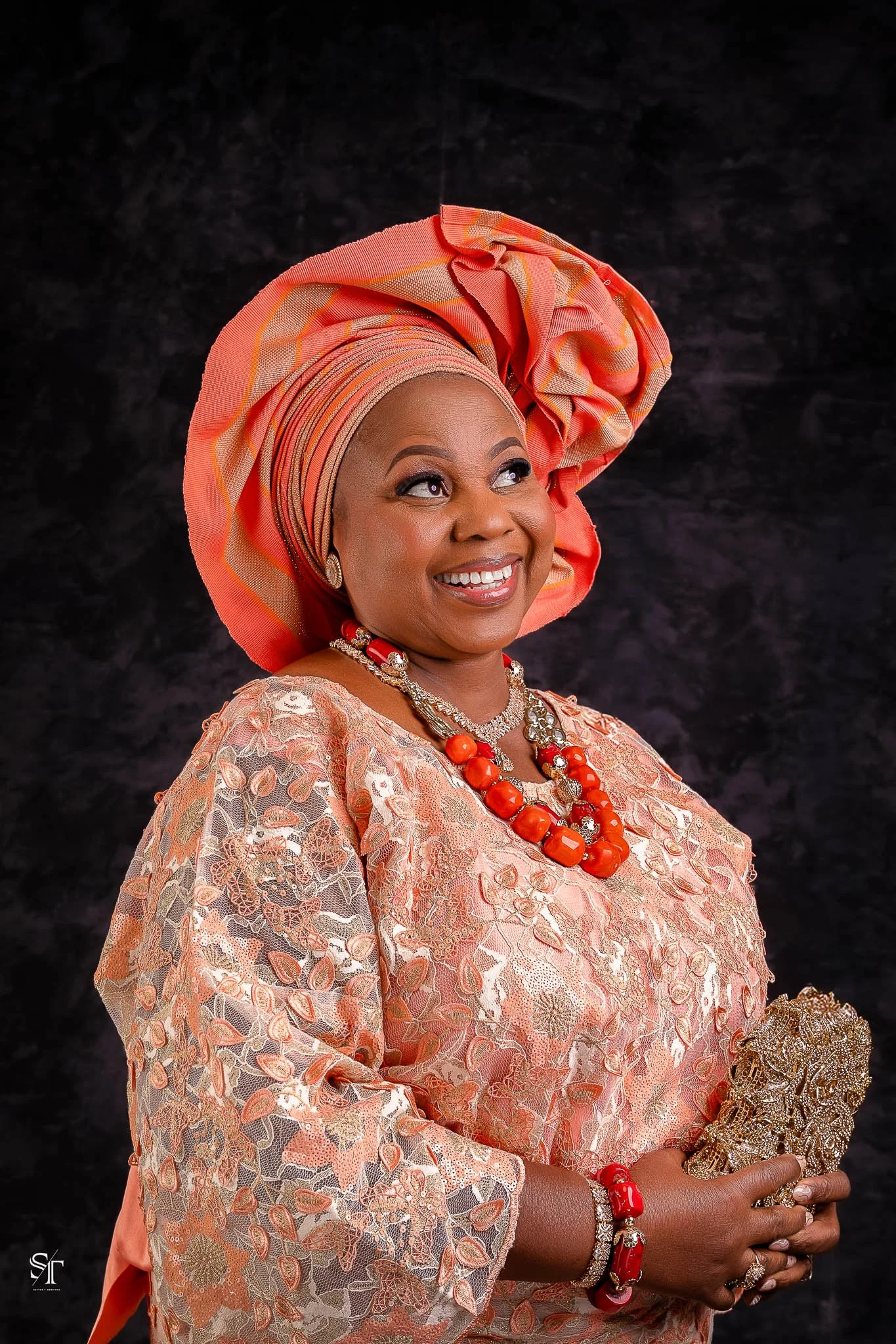 A woman dressed in traditional Nigerian attire with a coral headwrap, smiling, holding a silver accessory, against a dark background.