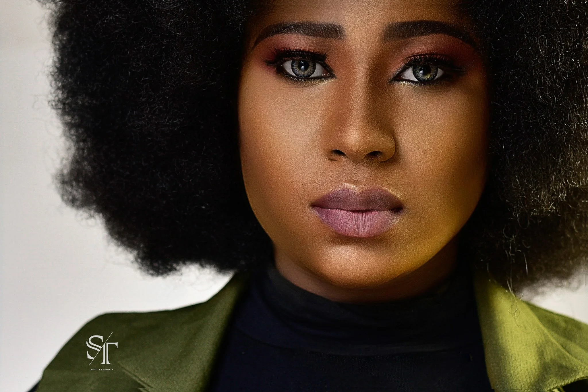 Close-up of a woman with dark skin, curly afro hair, and makeup, wearing a black turtleneck and an olive green jacket.