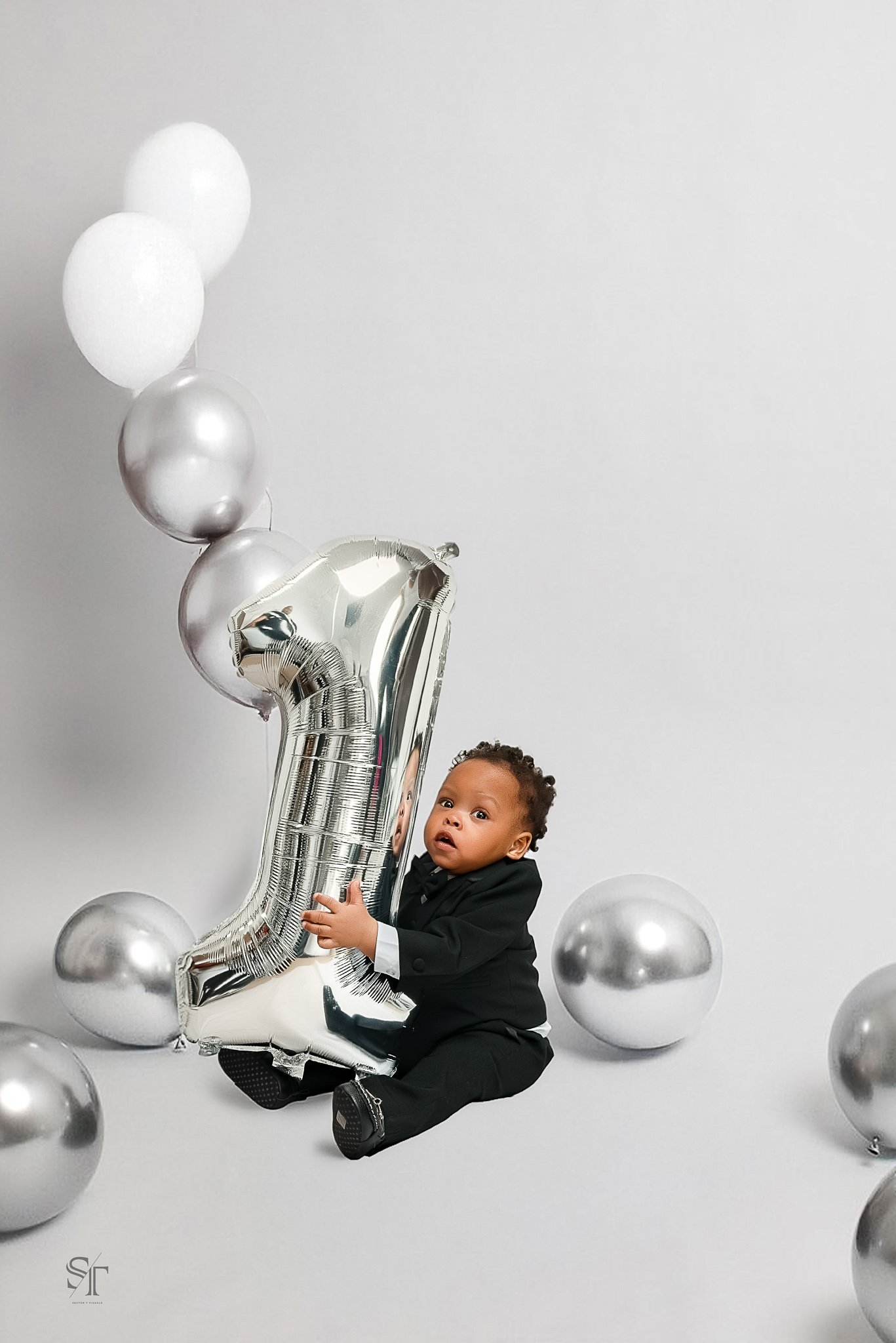 A young child dressed in a black suit sitting on the floor, holding a large silver number one balloon, surrounded by several silver and white balloons, against a plain white background.