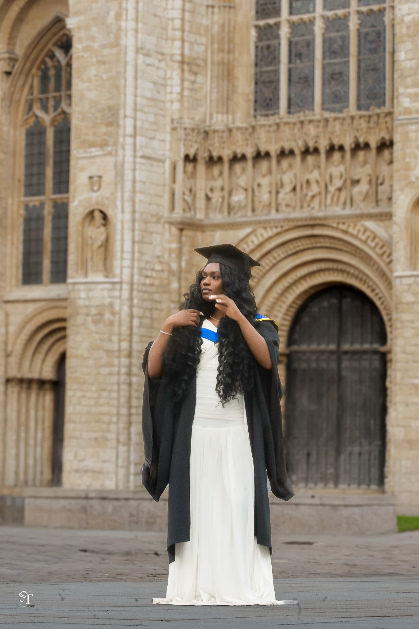 A young woman in a graduation gown and cap standing in front of a historic stone building with large arched windows and intricate carvings.