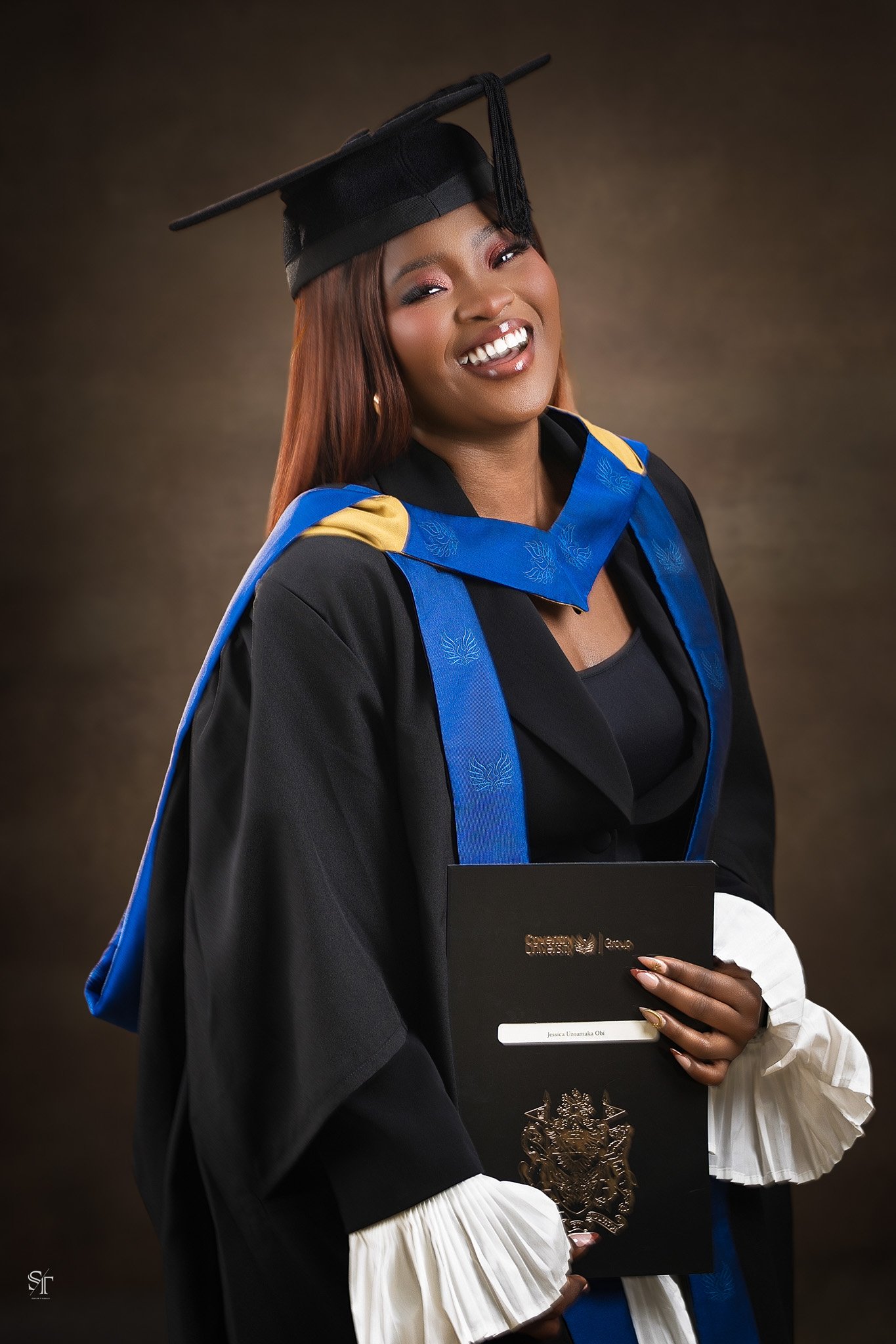 Woman in graduation gown and cap with blue and yellow hood, holding diploma against brown background.