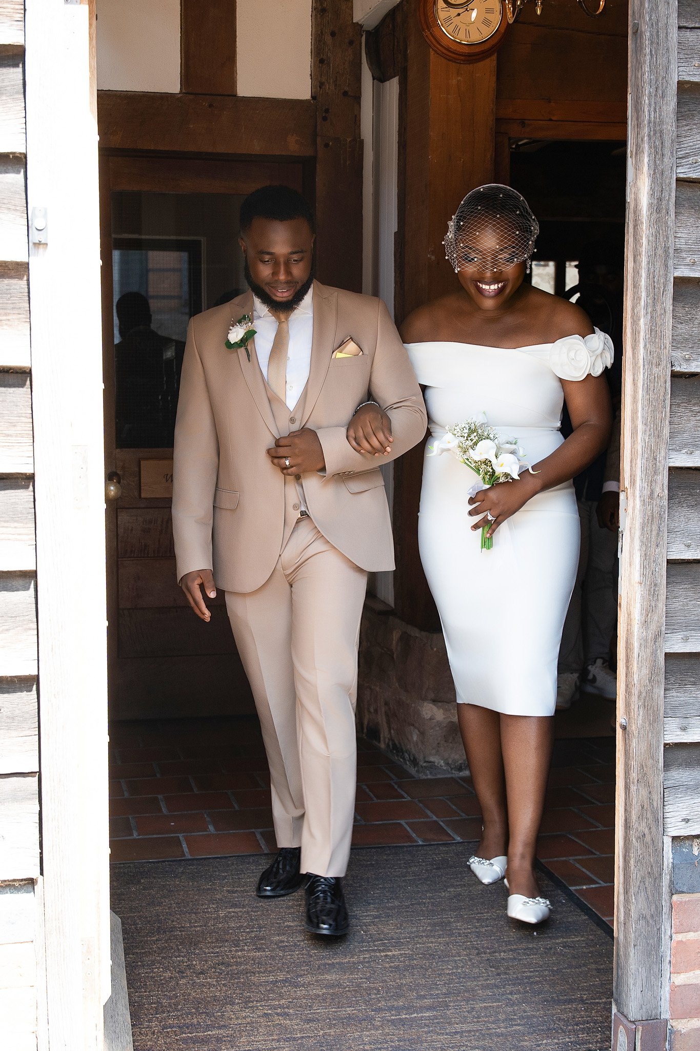 A bride and groom walking out of a building door. The groom wears a beige suit with a boutonniere, and the bride wears a white off-shoulder dress with flowers and a birdcage veil, holding a bouquet of white flowers. They are smiling and arm-in-arm.