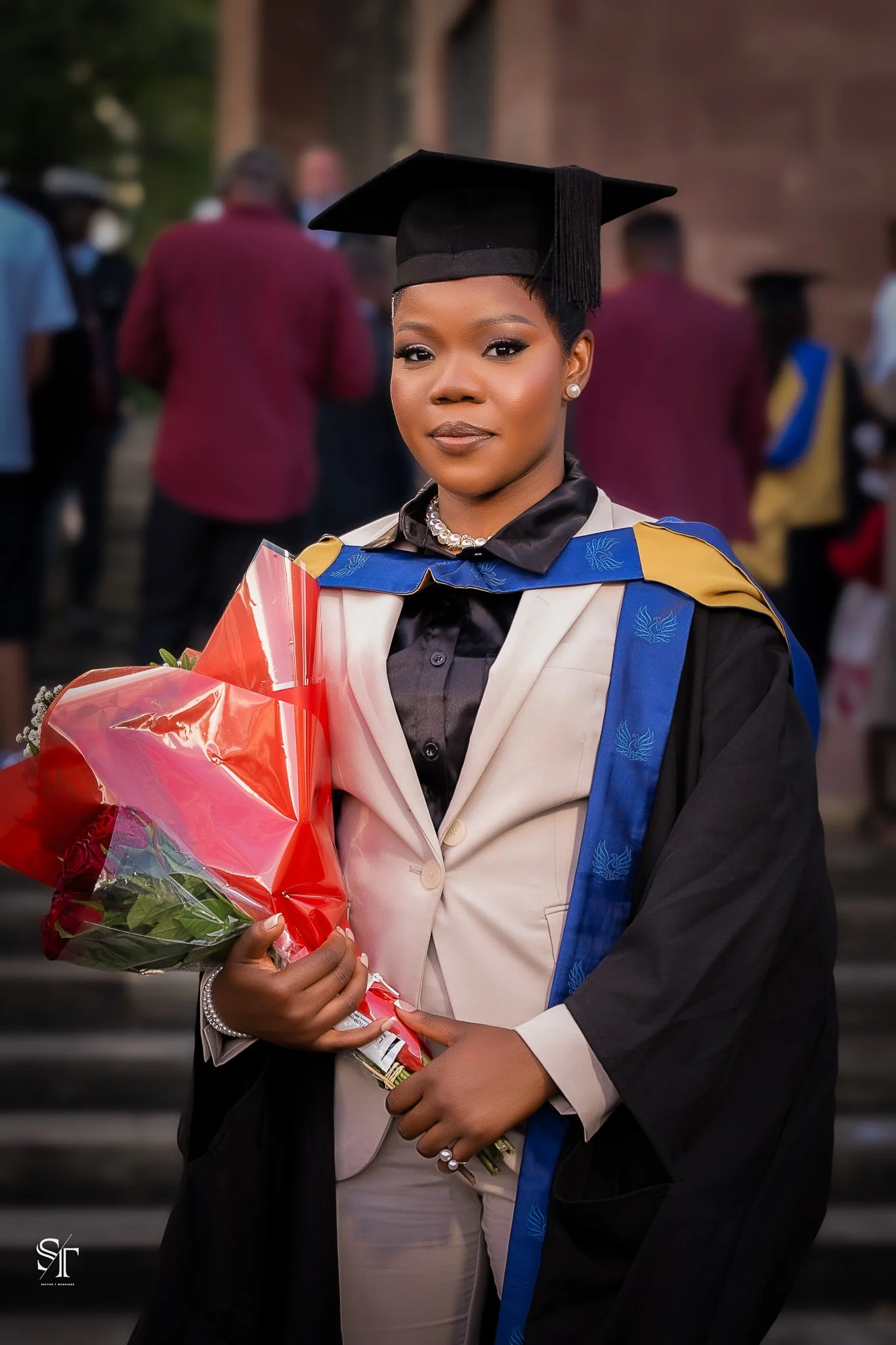 A woman in graduation attire holding a bouquet of flowers, standing outdoors with other graduates in the background.