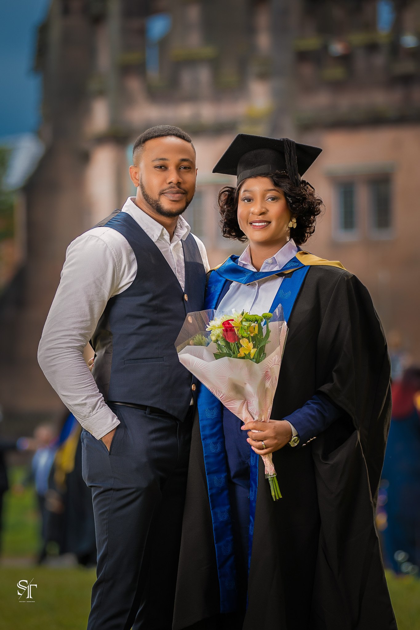 A young woman in graduation attire holding a bouquet of flowers standing next to a man in semi-formal clothing outdoors.