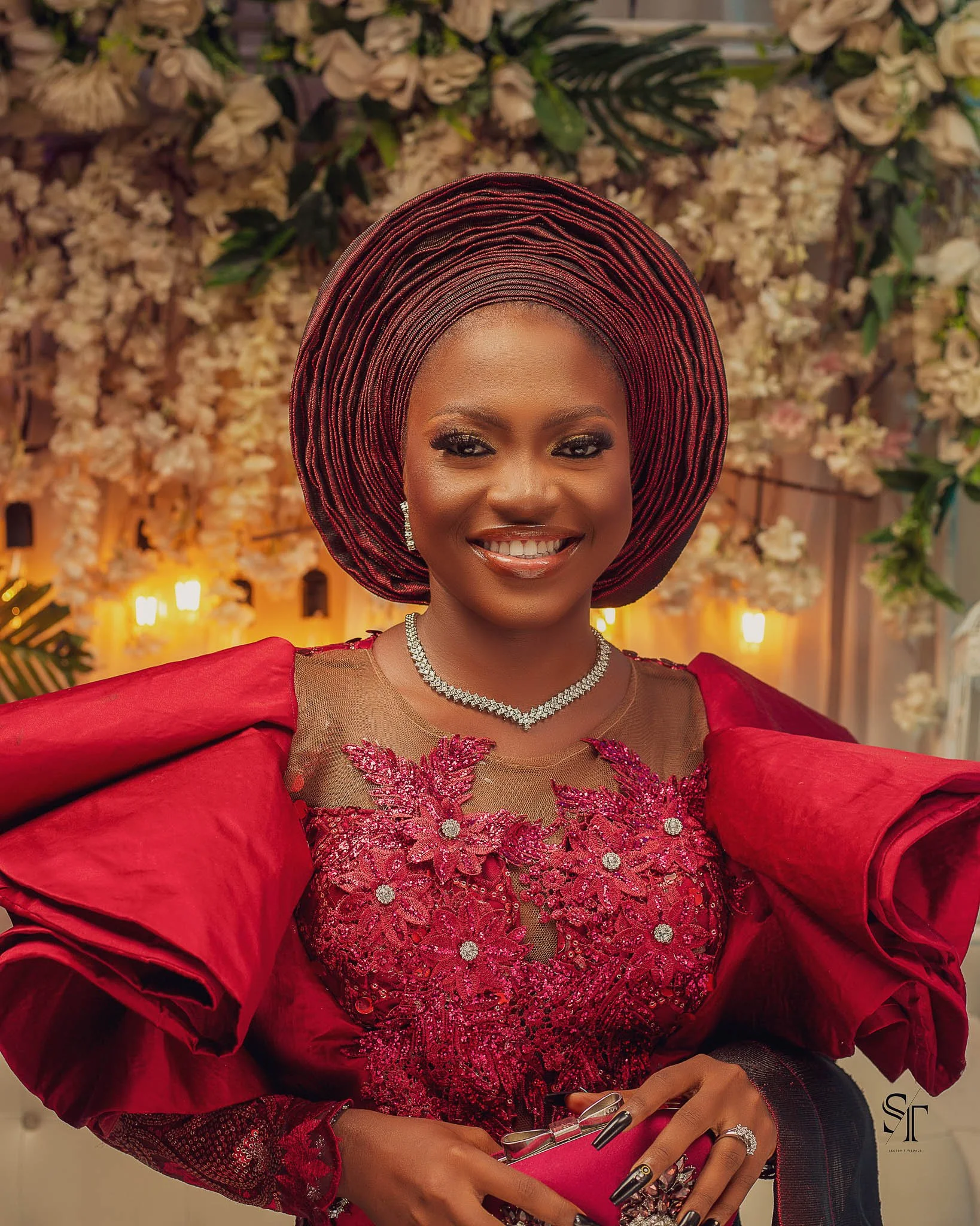 A woman dressed in a red lace gown with floral embroidery and puffed sleeves, wearing a large red and black gele, jewelry including a diamond necklace, earrings, and ring, standing in front of a floral backdrop with warm lighting