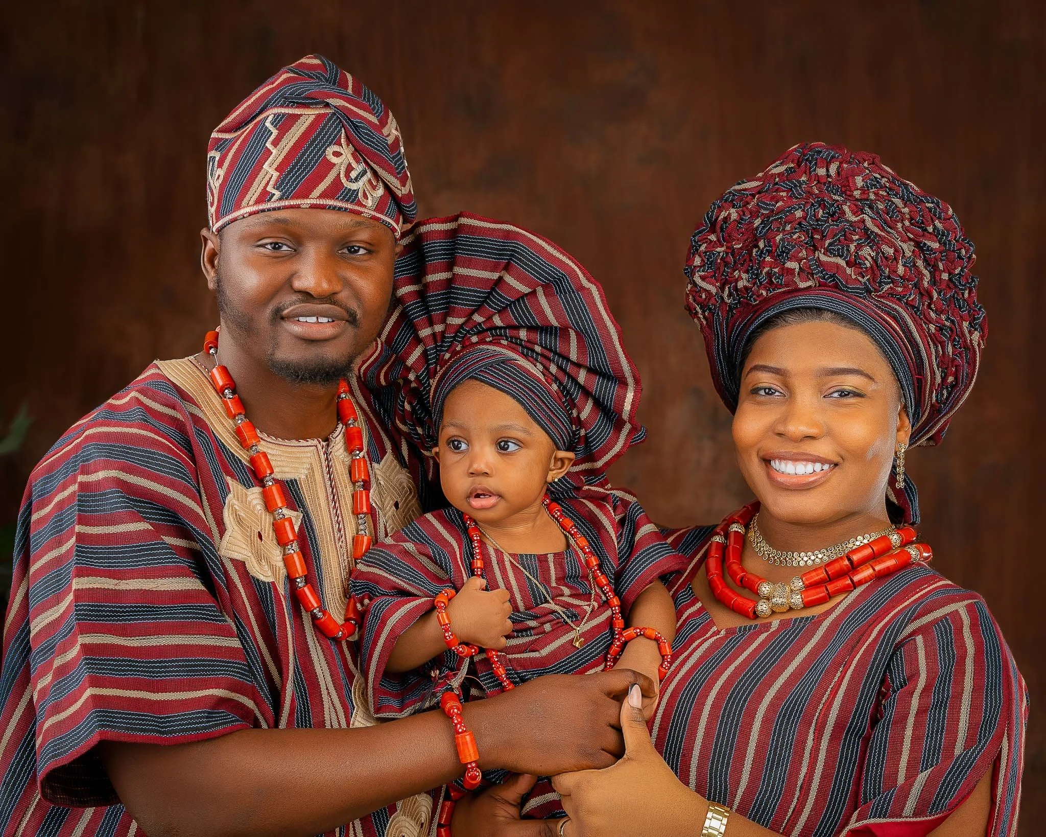 A family of three dressed in traditional African attire, with the father and mother holding their young child in front of a brown backdrop.
