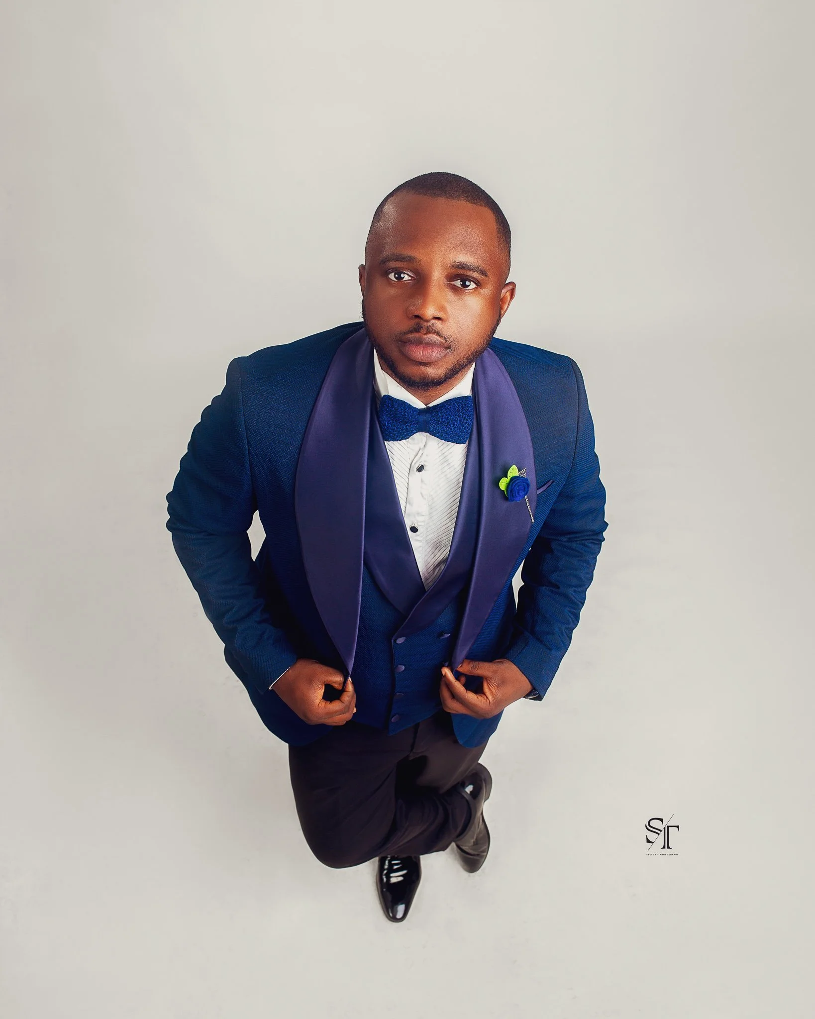 A man in a blue tuxedo with a bow tie and white shirt posing against a plain background, looking directly at the camera.