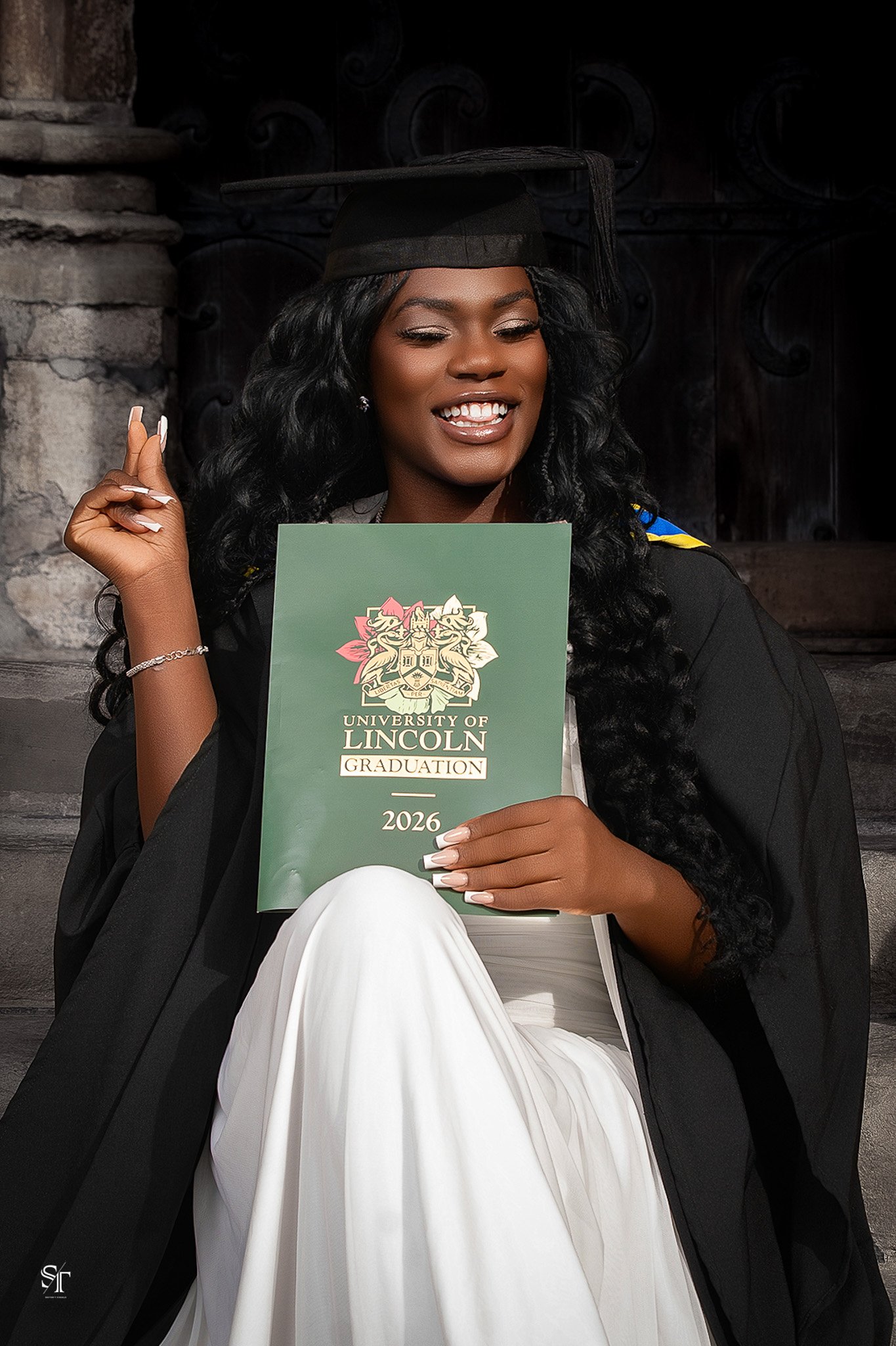 Young woman in graduation cap and gown holding a University of Lincoln diploma. She has long curly black hair, is smiling, and seated in front of a stone wall.