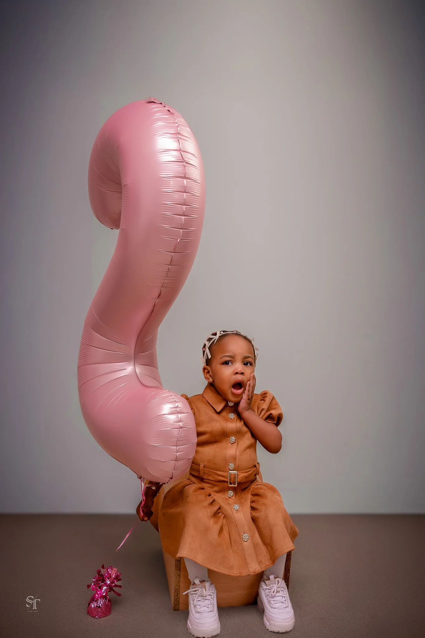 A young girl sitting on a small wooden chair, holding a large pink heart-shaped balloon with a surprised or shocked expression on her face. The girl is wearing a brown dress with puffy sleeves, white sneakers, and a headband with a bow. The backgroun