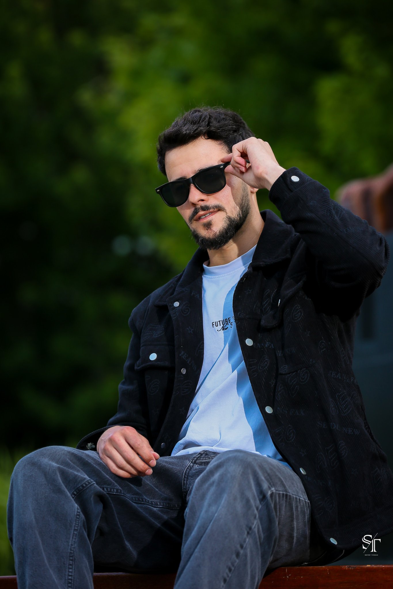 A young man with dark hair, a beard, and sunglasses sitting outdoors on a park bench, adjusting his sunglasses with his right hand, with a blurred green background.