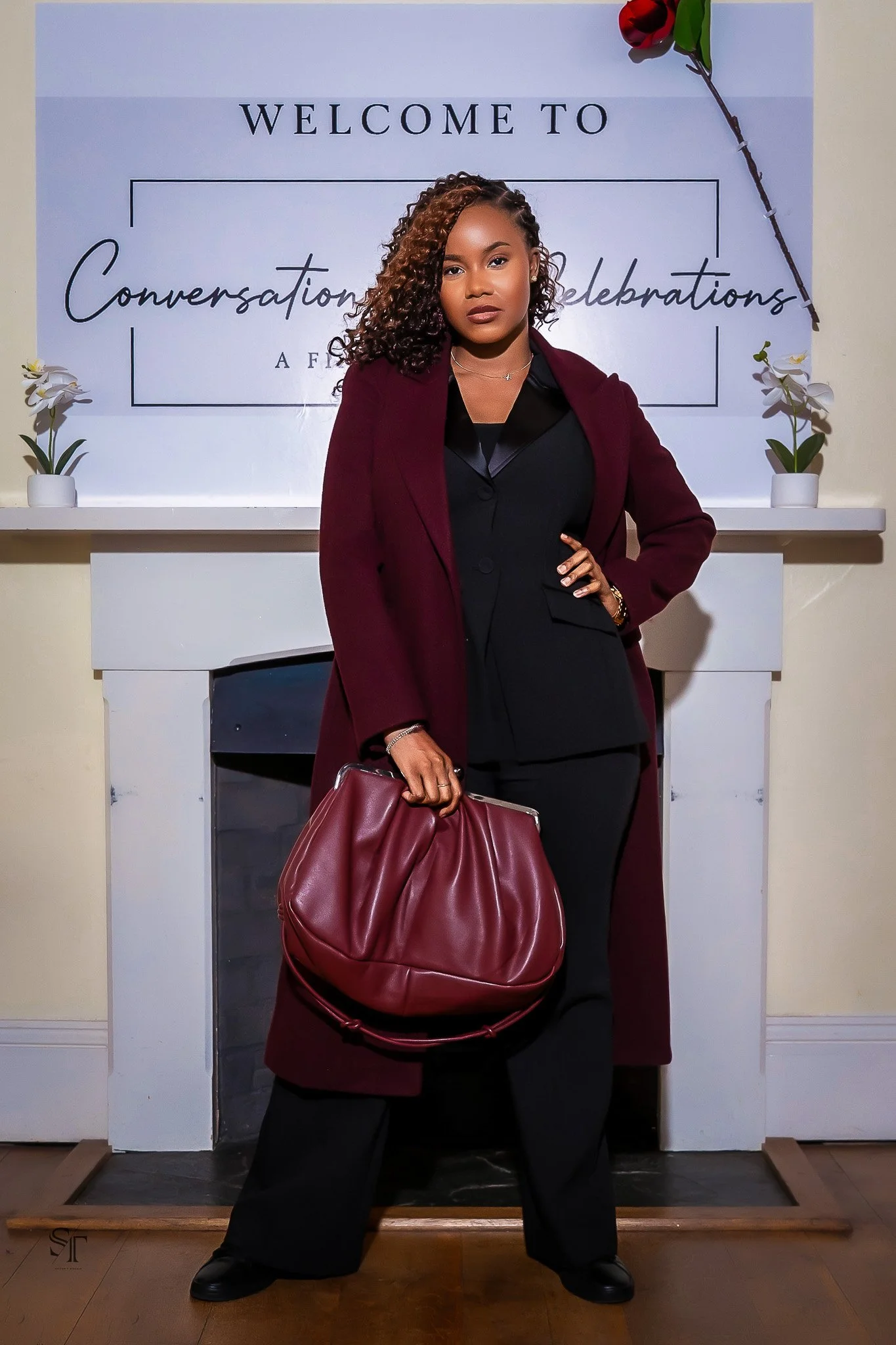 A woman standing in front of a welcome sign for a celebration event, holding a large red handbag, wearing a black outfit and a burgundy coat.