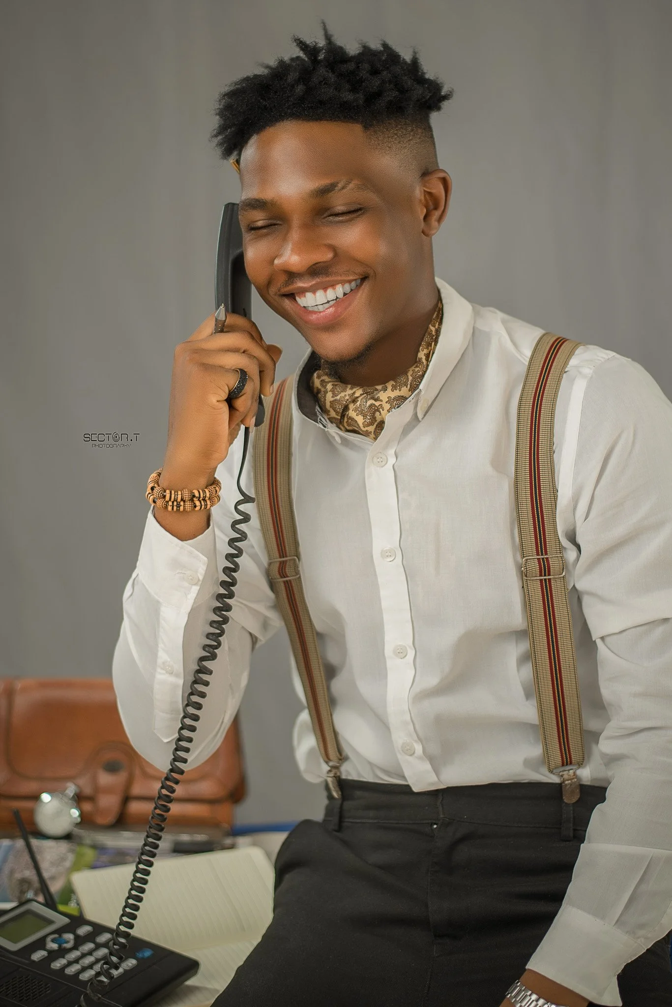 Young man smiling while talking on a corded telephone, dressed in a white shirt with suspenders and a patterned scarf, with a watch and beads on his wrist, in an office setting.