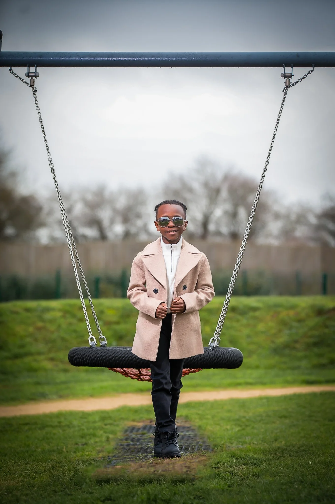 A young boy with sunglasses and a beige coat standing on a black net swing in a park.