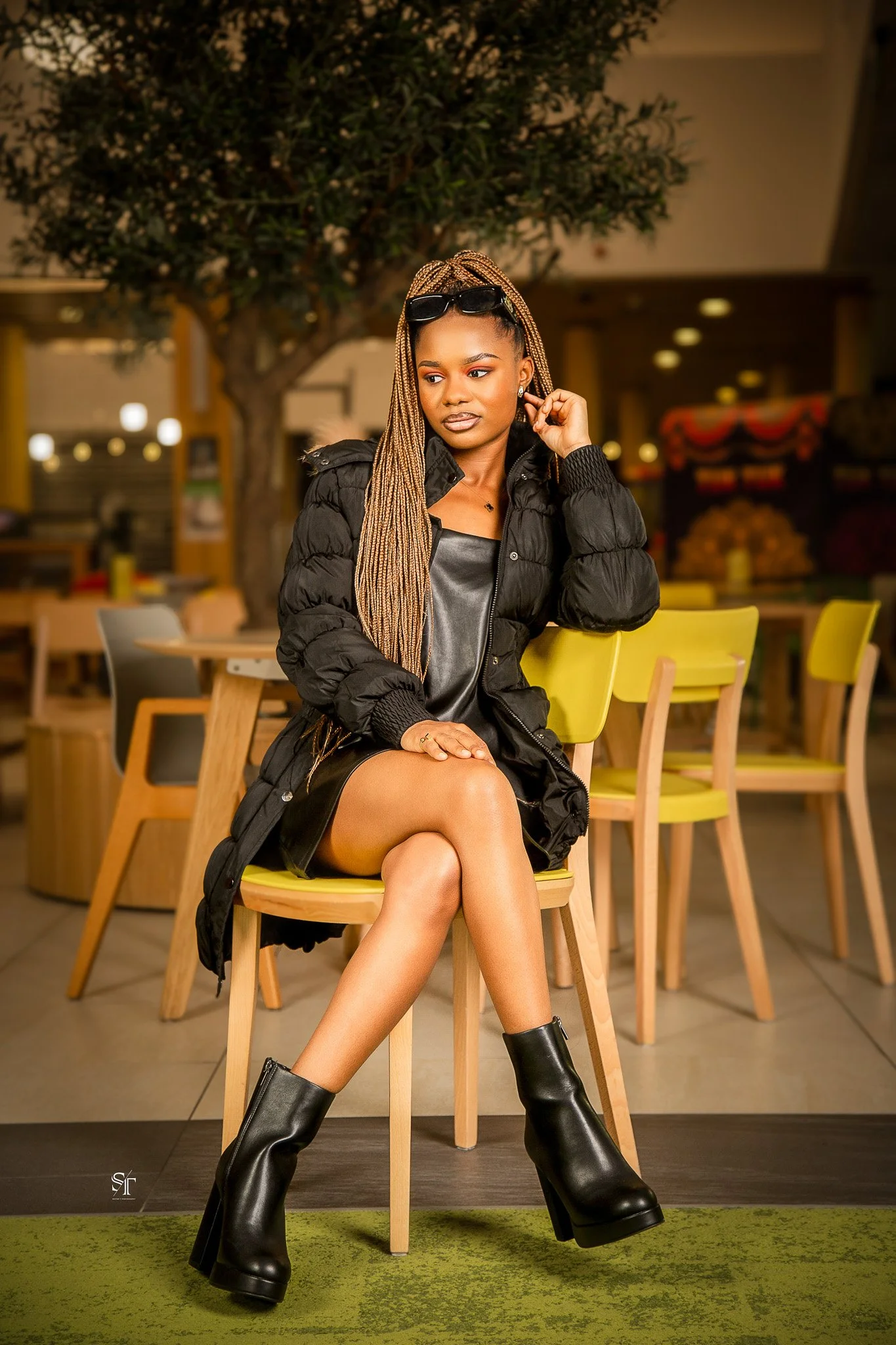 A stylish woman sitting on a yellow chair in a cafe or restaurant. She has long braids, is wearing sunglasses on her head, a black puffer jacket, a black leather dress, and black high-heeled boots. She is sitting with one leg crossed over the other.