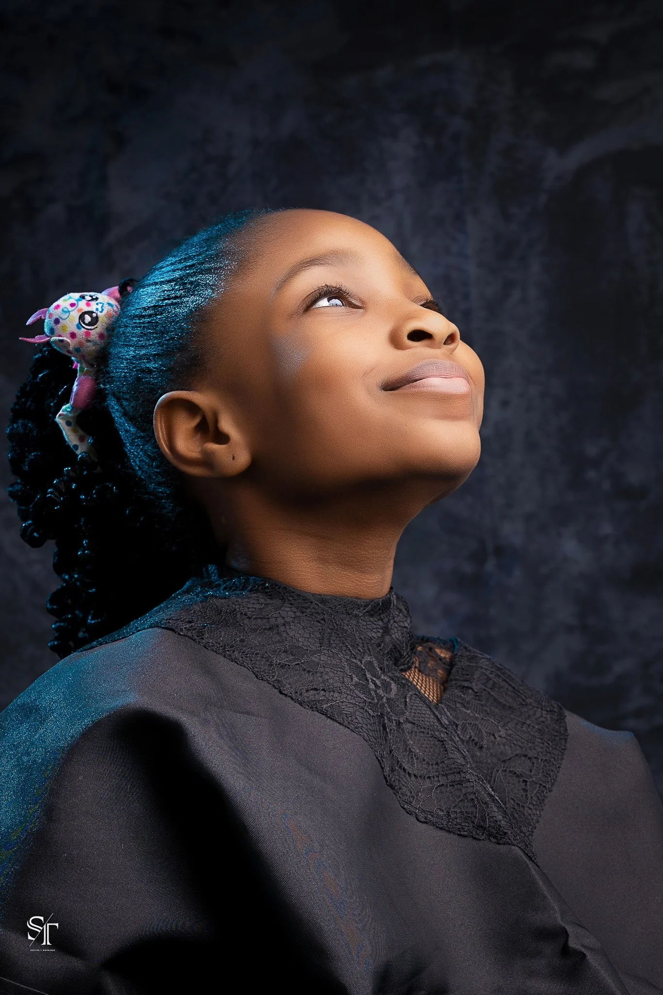 A young girl with a joyful expression, looking upward, wearing a black lace dress and a colorful hair bow, against a dark background.