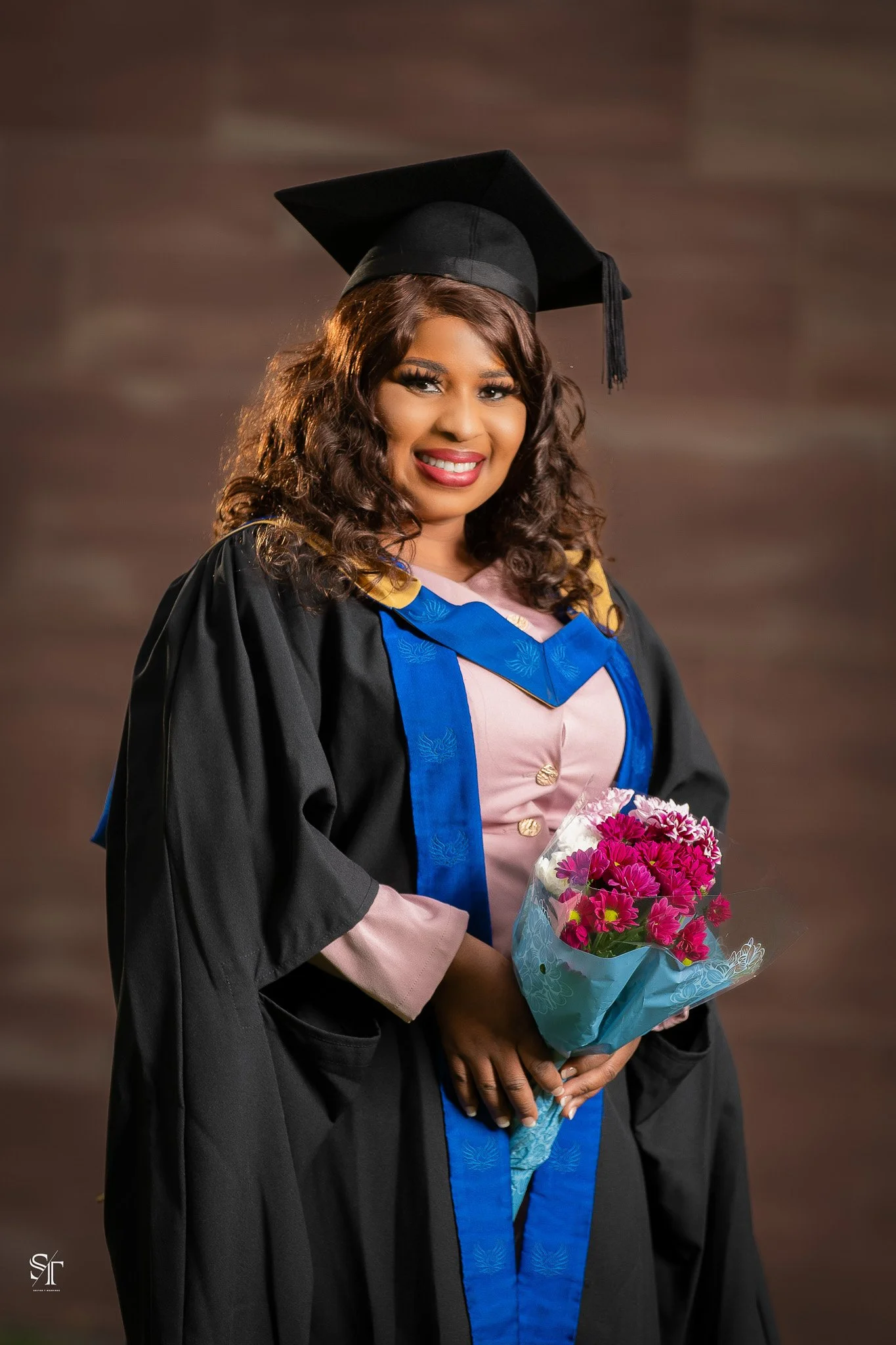 A woman wearing a black graduation cap and gown holding a bouquet of pink flowers, smiling in a studio with a brown background.