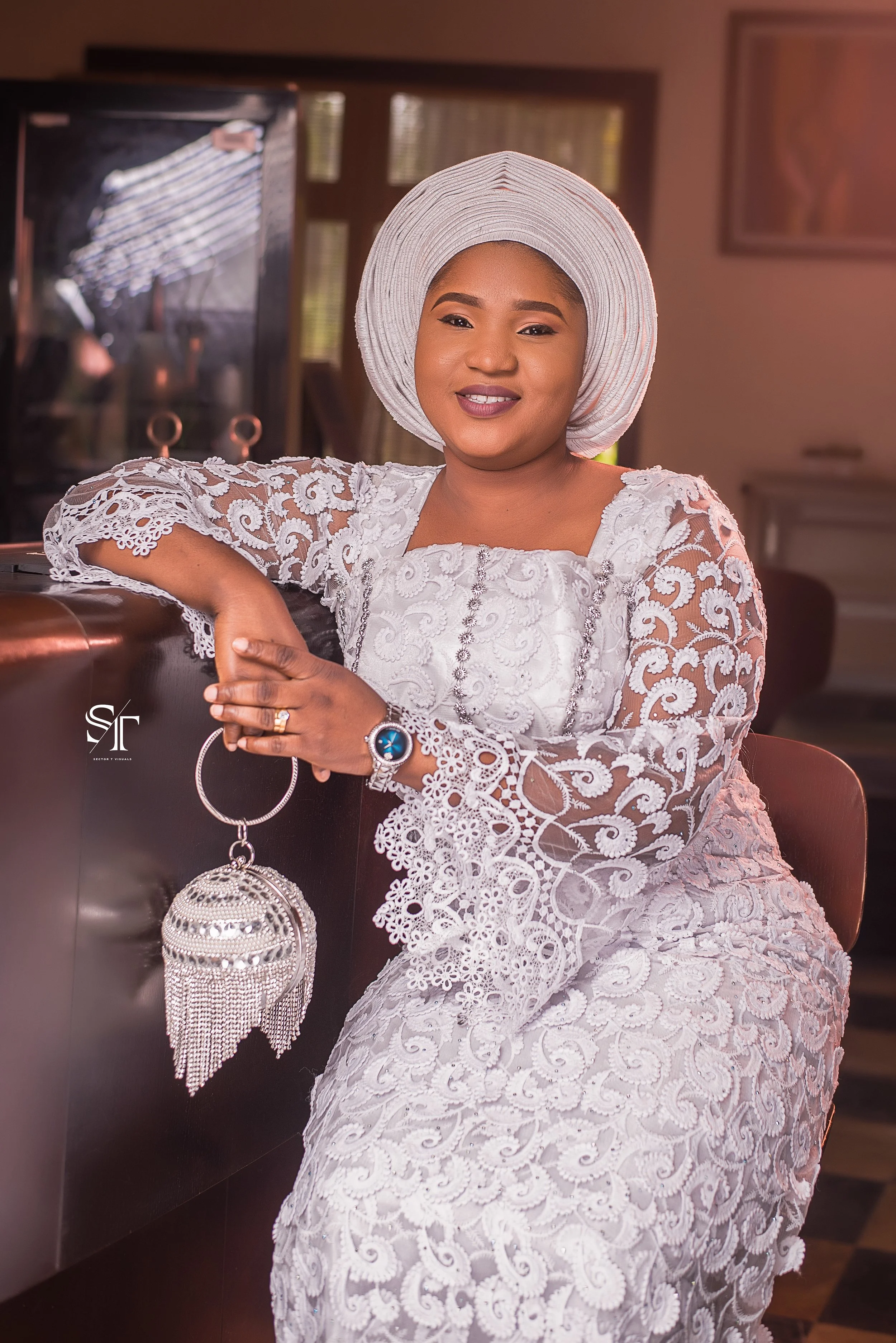 A woman dressed in an elegant white lace outfit with intricate embroidery, sitting indoors with her arm resting on a shiny surface. She is wearing a matching wide-brimmed headwrap, a watch, and a ring, and is smiling at the camera.