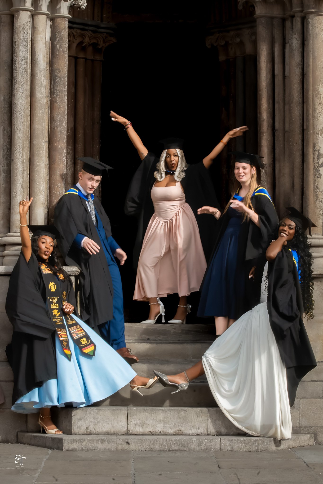 Group of seven young women in graduation caps and gowns posing on the steps of a historic building with columns.