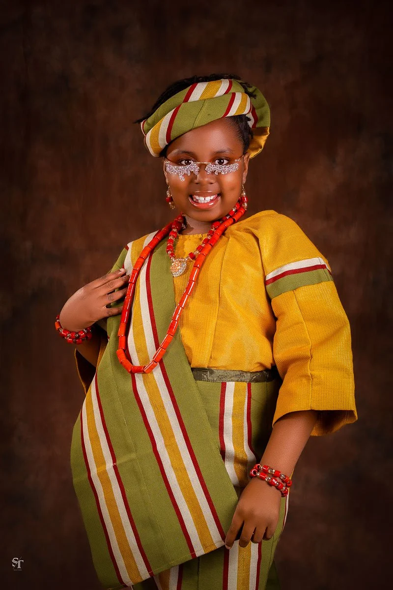 A young girl wearing traditional African attire with colorful beaded jewelry and a matching headwrap, posing against a dark background.