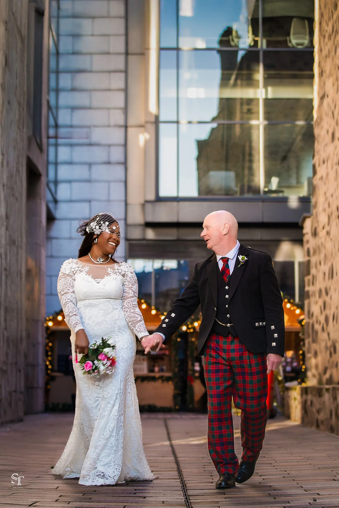 Bride and groom walking hand in hand, smiling, outside at a city wedding, with modern glass building background and warm evening lighting.