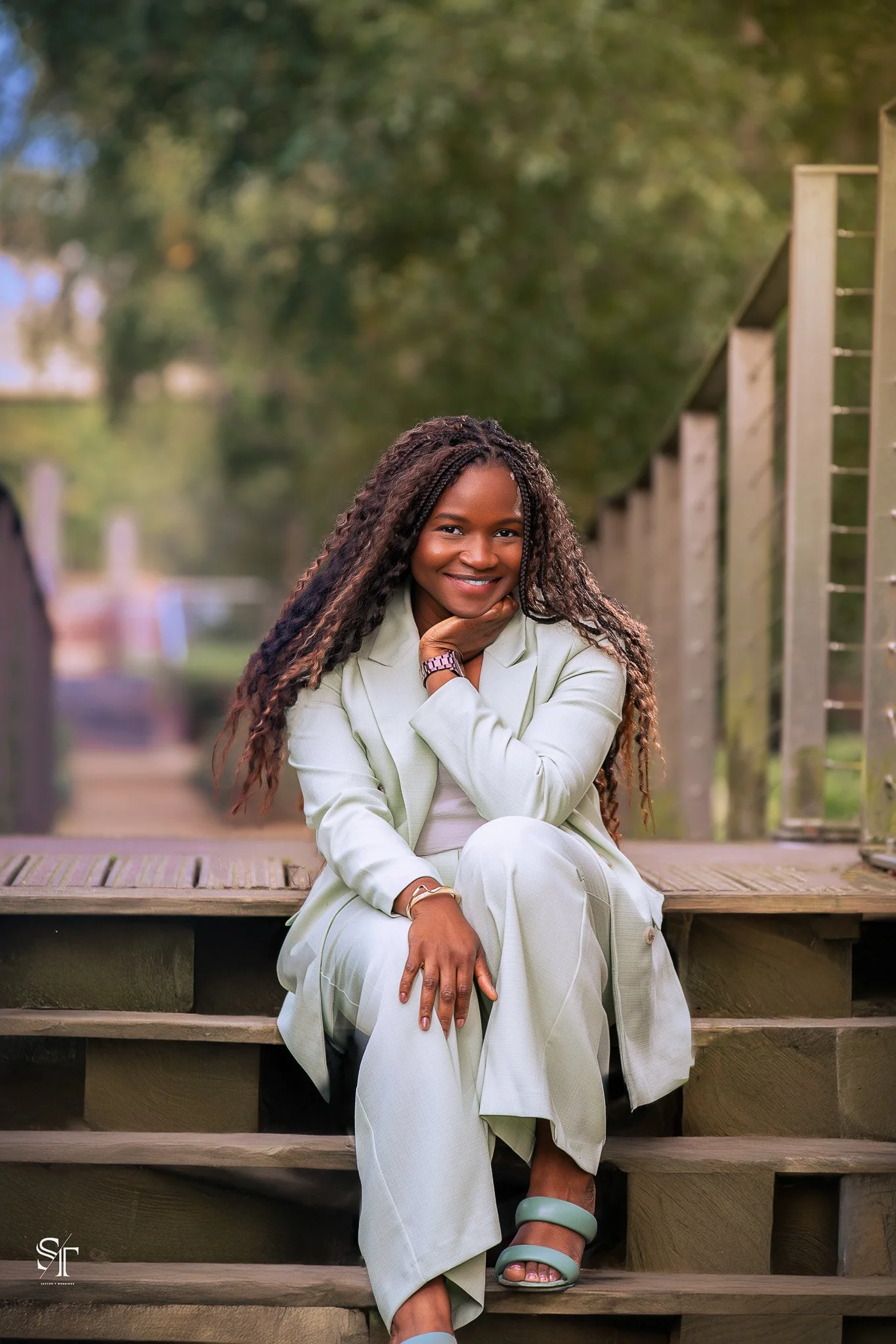 A woman sitting on wooden steps outdoors, smiling, wearing a light-colored suit, with long curly hair, with trees in the background.