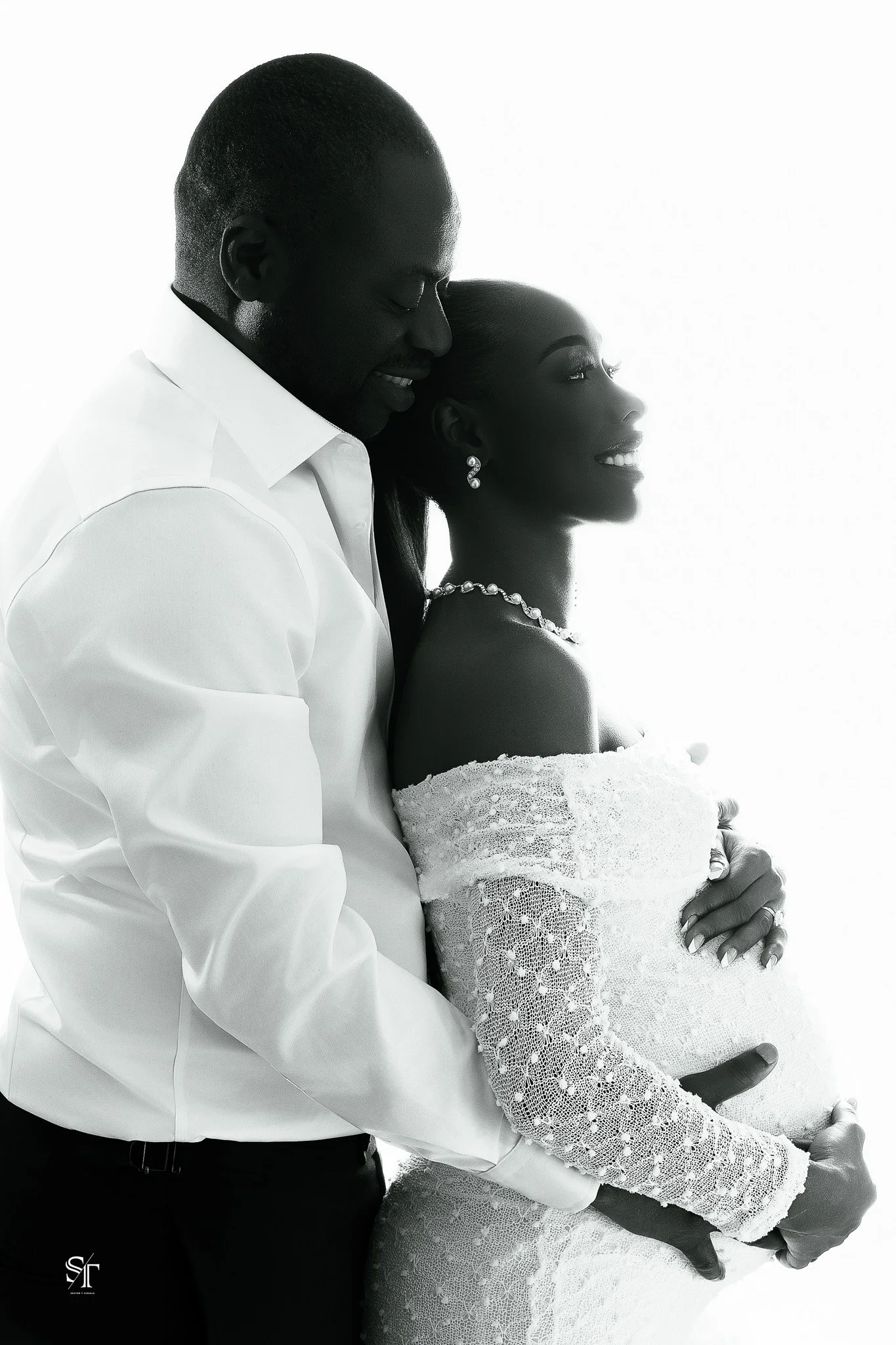 A black-and-white photograph of a man and woman in wedding attire. The man is wearing a white shirt and the woman is in a lace wedding dress with jewelry. They are standing close together, sharing an intimate moment with their foreheads touching and 