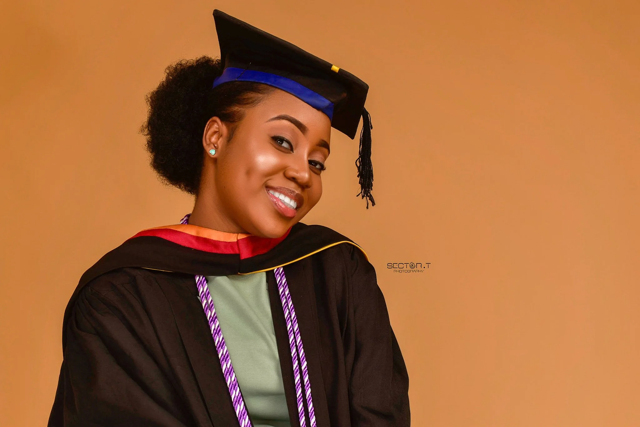 A young woman with curly hair wearing a graduation cap and gown, smiling against a plain background.
