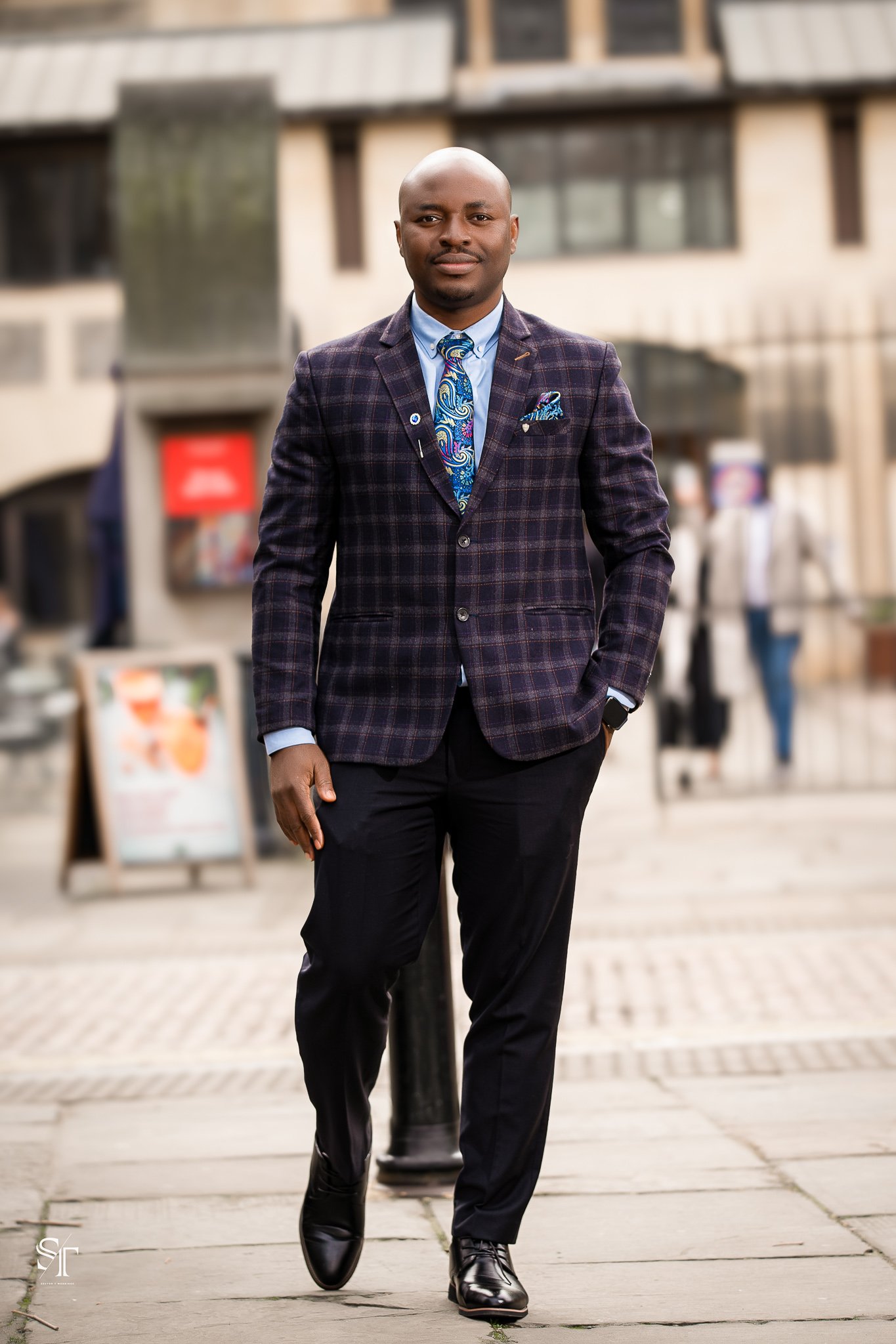 A man walking outdoors in business attire, wearing a plaid blazer, light blue shirt, patterned tie, black pants, and black leather shoes.
