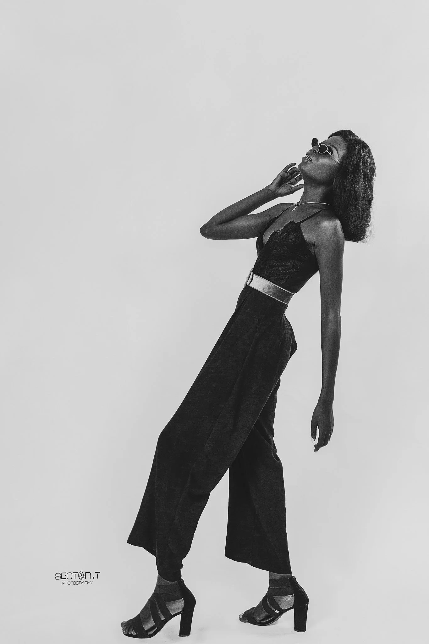 A black and white photo of a fashionable woman wearing sunglasses, a lace top, high-waisted pants, and high heels, striking a pose against a plain background.