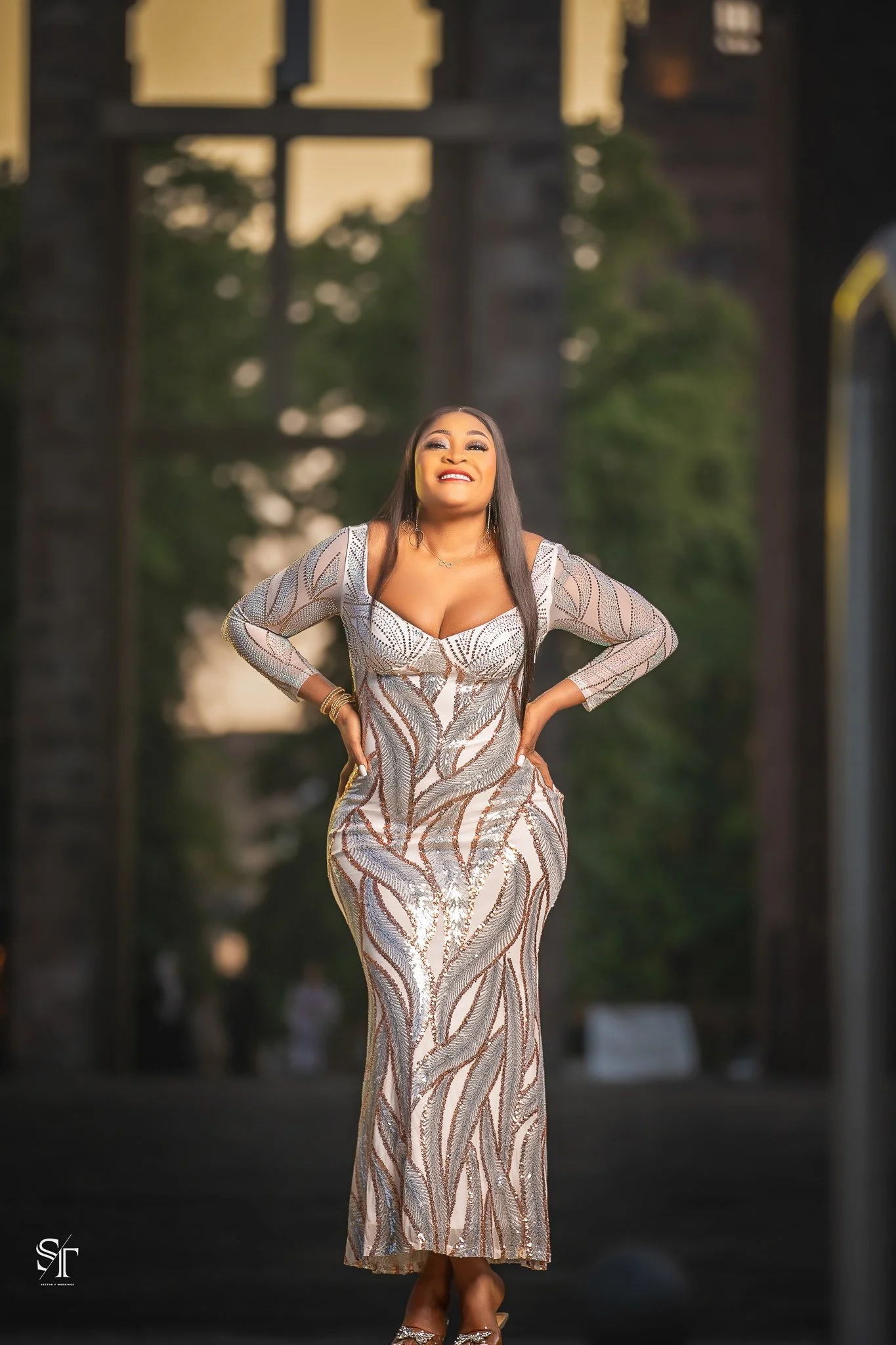 A woman wearing an elegant, sequined gown with intricate silver and tan patterns, standing outdoors with her hands on her hips and smiling confidently.