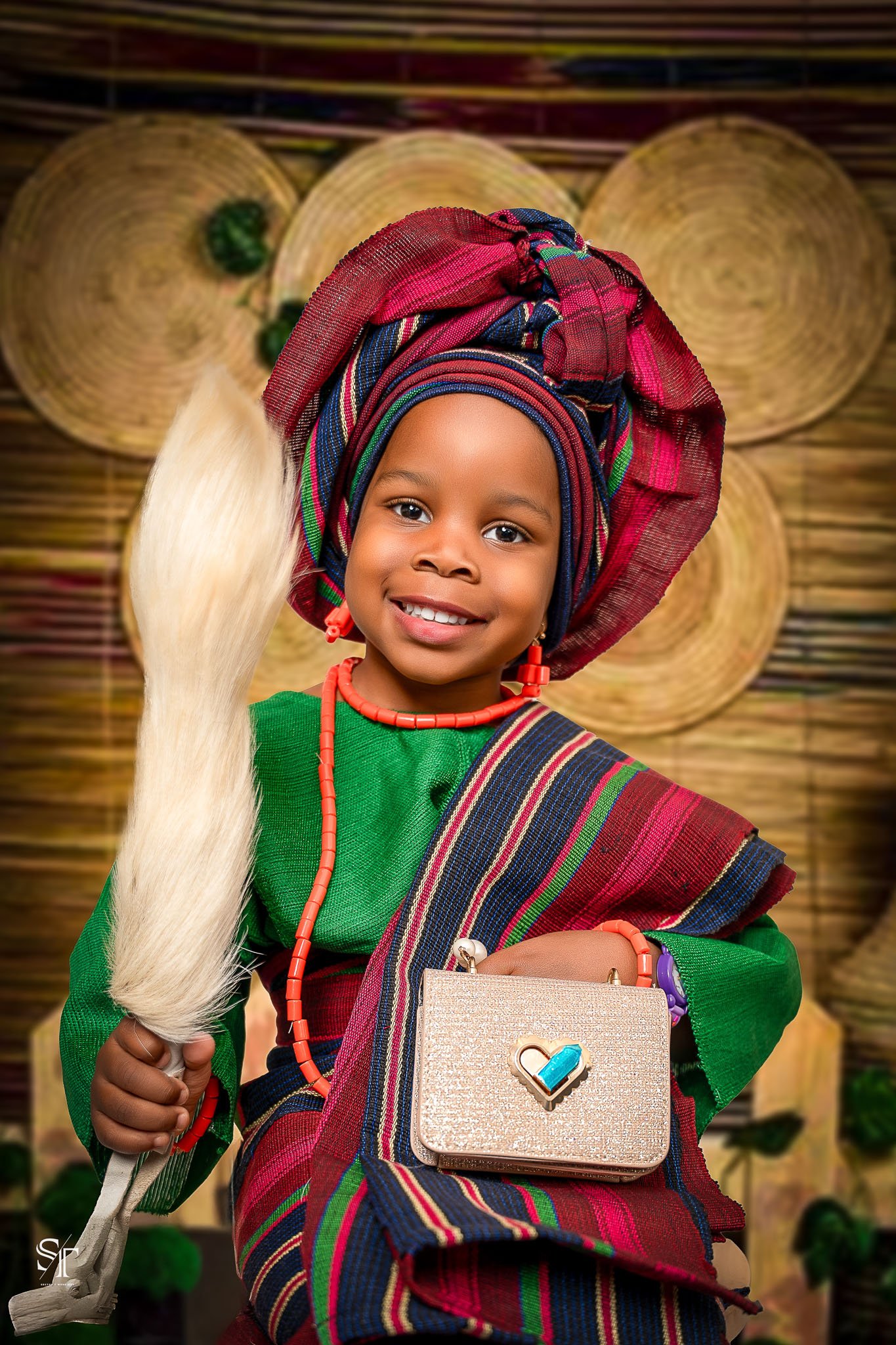 A young girl dressed in traditional African attire, wearing a colorful headwrap, necklace, and earrings, holding a feather and standing in front of a wooden background with woven baskets.