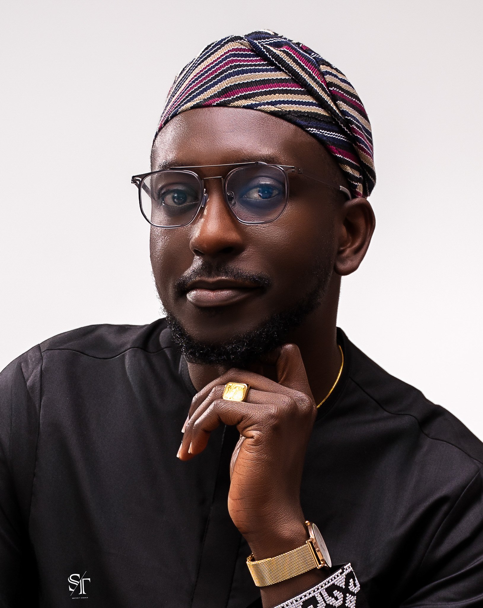 A portrait of a man wearing glasses, a colorful head wrap, a black shirt, and gold accessories, posing with his chin resting on his hand.