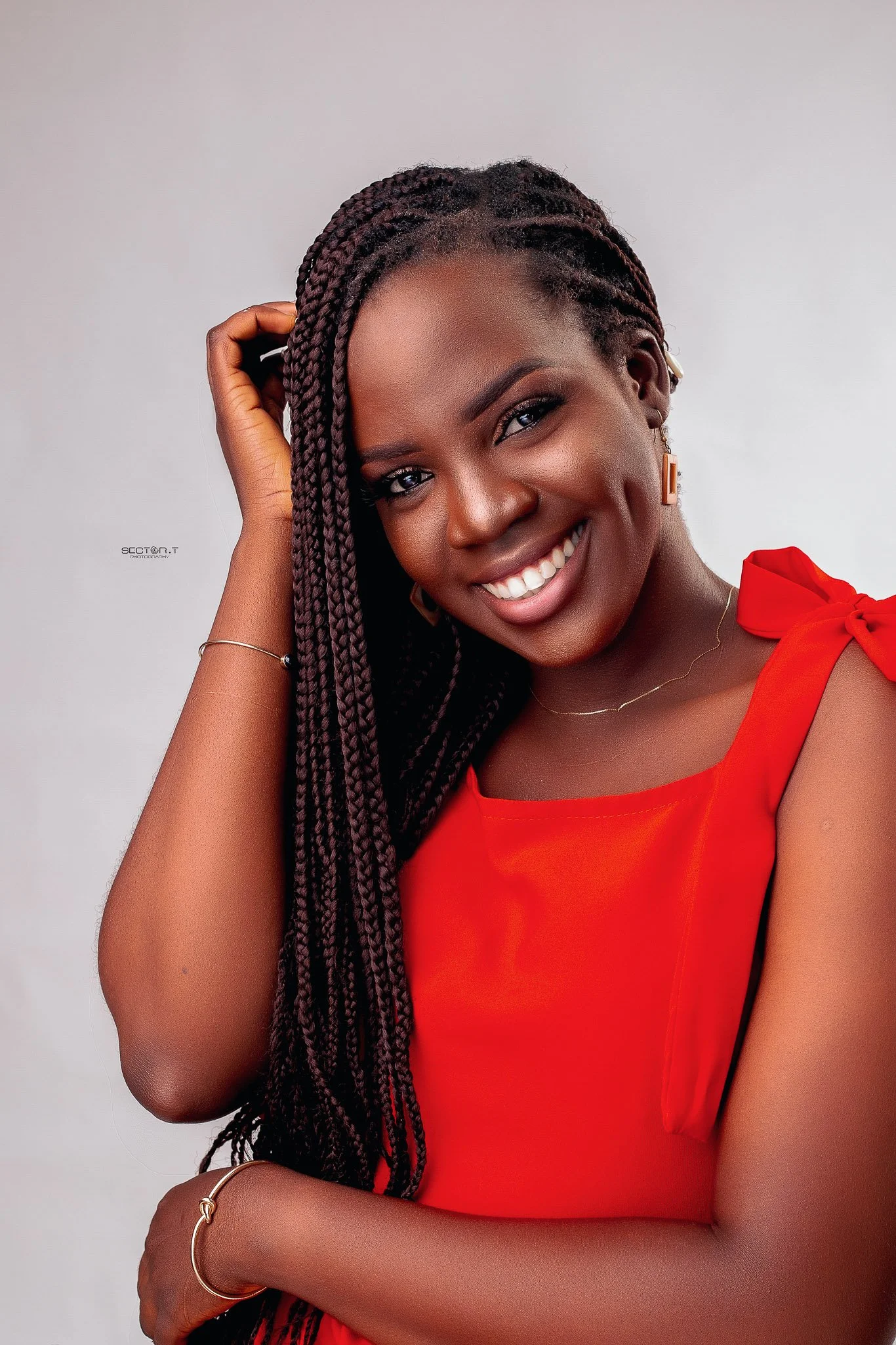 A young woman with dark skin, styled with long braided hair, smiling thoughtfully, wearing a red top with shoulder bow, minimal jewelry, against a simple light background.