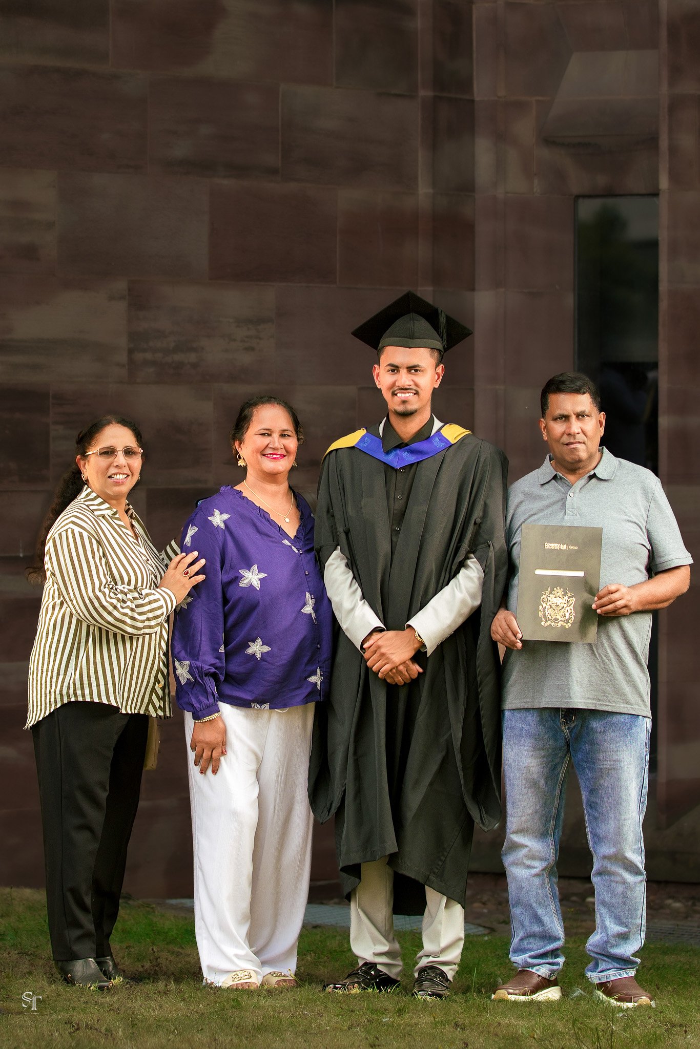 A young man in a graduation gown and cap standing with four family members in front of a dark wall, celebrating his graduation.