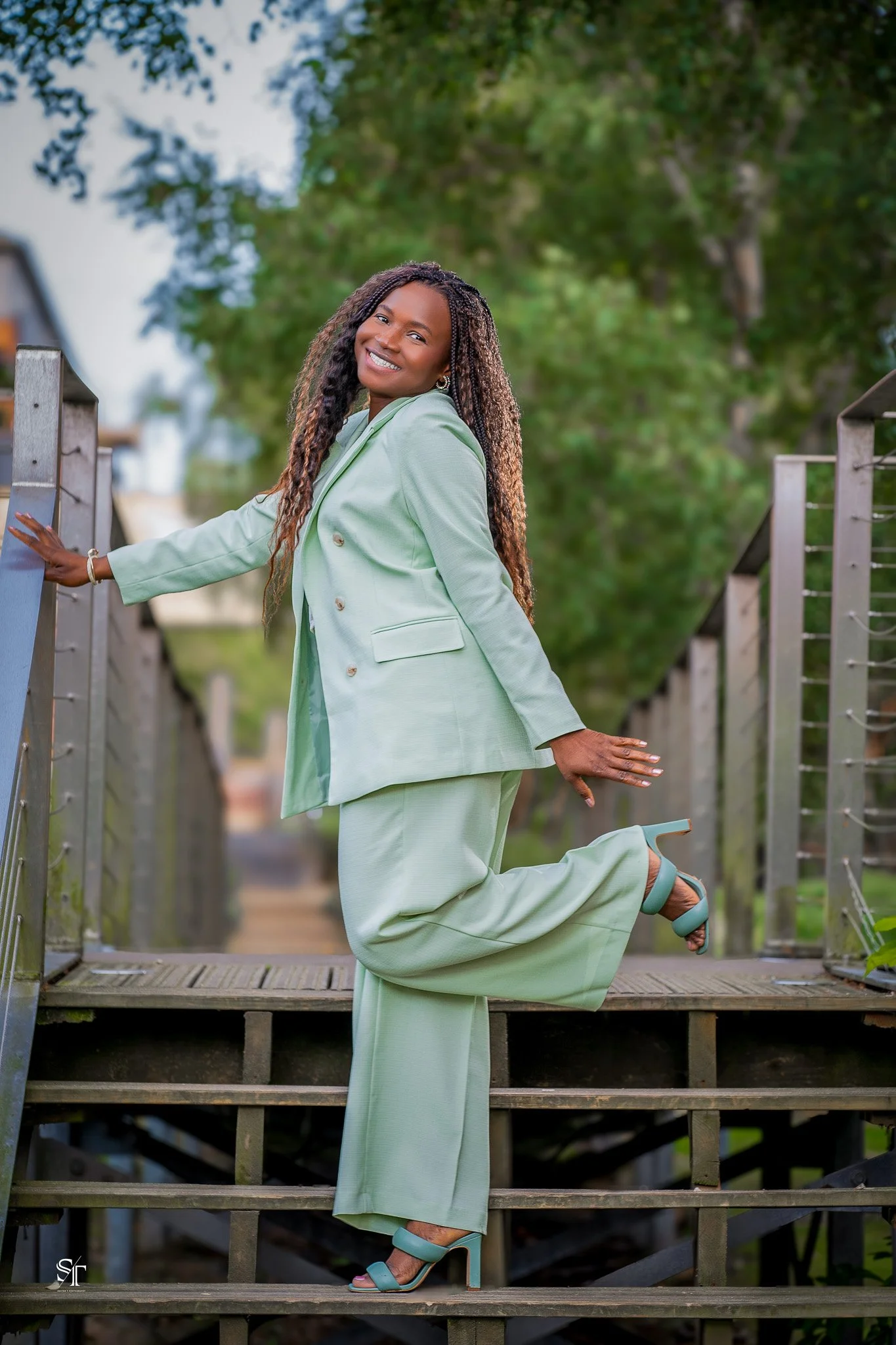 A woman dressed in a mint green pantsuit with open-toe heels, standing on a wooden staircase outdoors with green trees in the background, smiling at the camera.