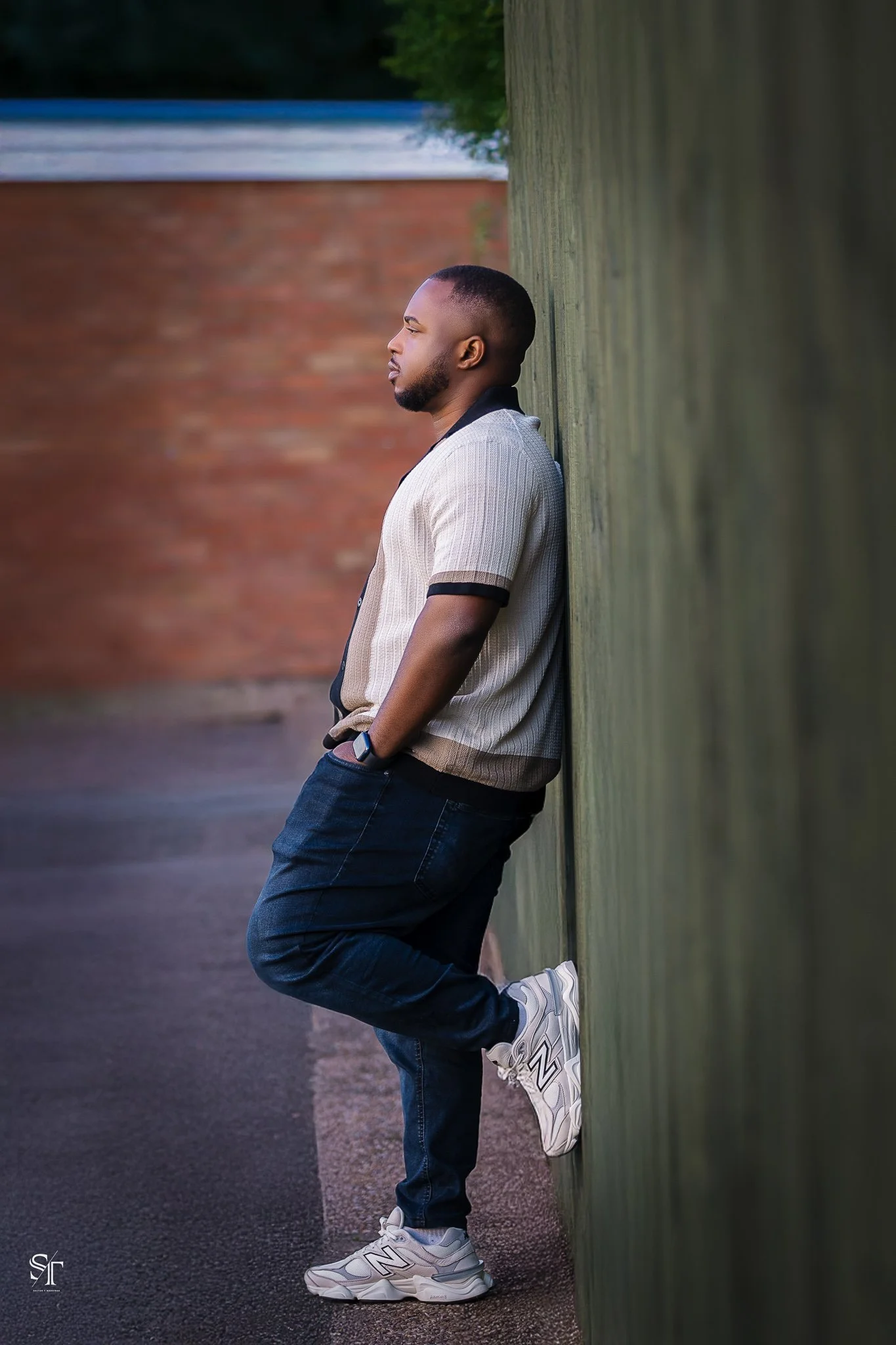 A man leaning against a green wooden wall outdoors, with arms in pockets, wearing a beige and black shirt, dark jeans, and white sneakers, looking to the left.