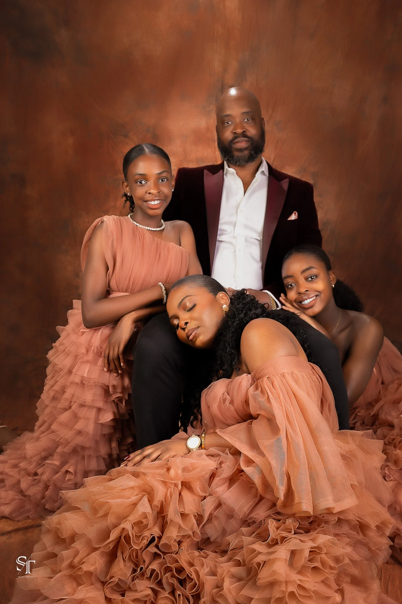 A family of five posing together in elegant, peach-colored dresses and a tuxedo. The woman in the front is sleeping on the man's lap, with all members smiling or peacefully resting against each other, against a brown background.