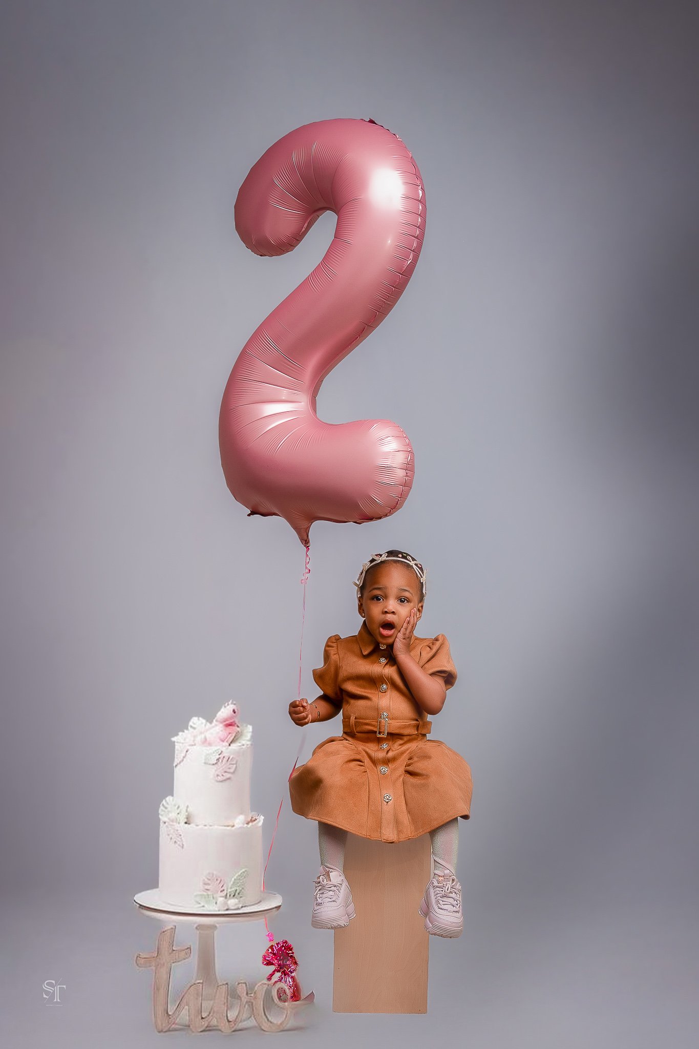 A young girl celebrating her second birthday with a large pink balloon in the shape of the number 2, a cake decorated with butterflies, and a "two" sign.