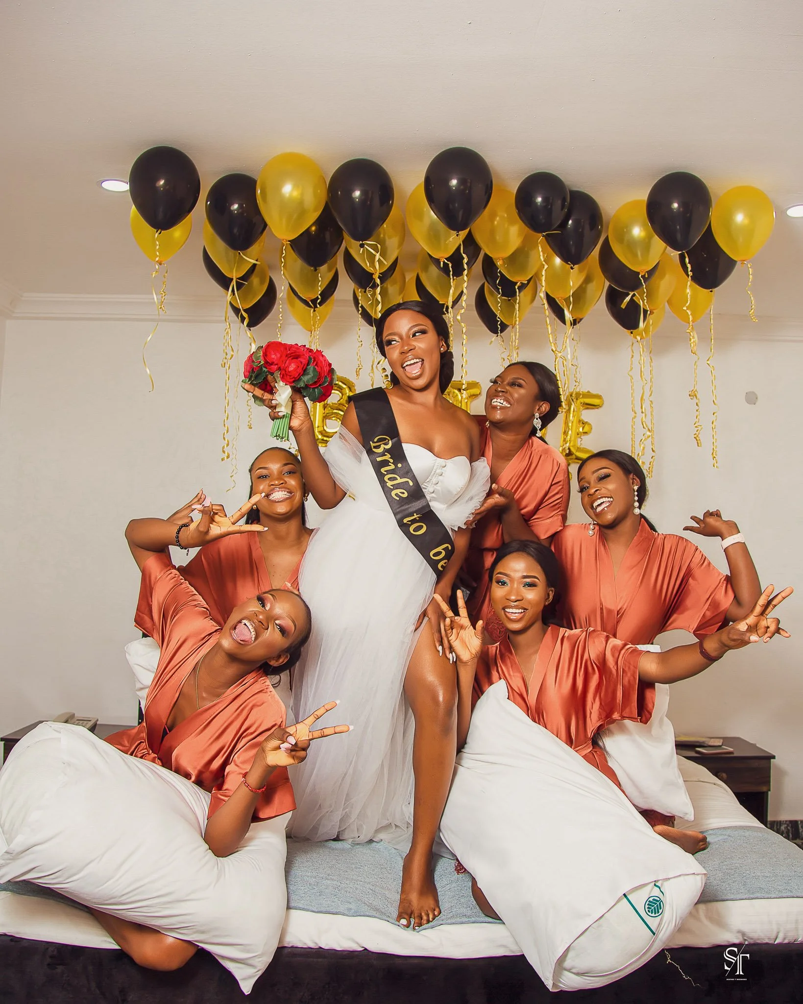 A bride in a white wedding dress with a black sash that says "Bride to be" stands on a bed surrounded by five women in matching peach-colored satin robes, all smiling and making peace signs. They are indoors with black and gold balloons behind them, 