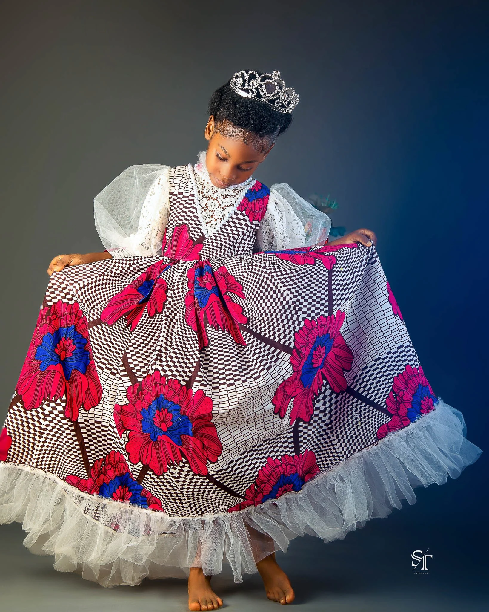 A young girl dressed in a vibrant African dress with pink and blue floral patterns, wearing a silver tiara, and holding the hem of her dress while standing barefoot against a plain background.