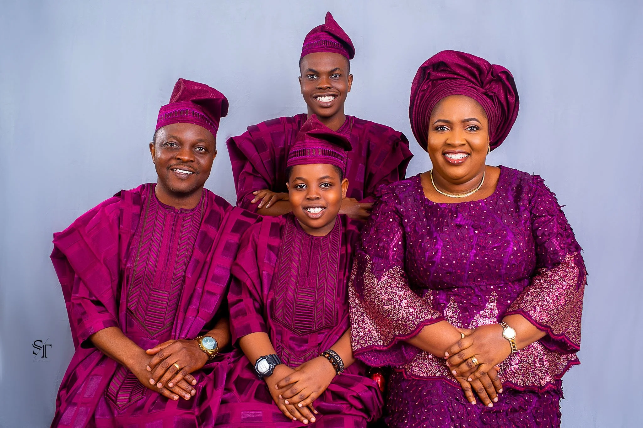 A Nigerian family dressed in traditional purple outfits, including two men, one woman, and two children, posing together against a plain background.
