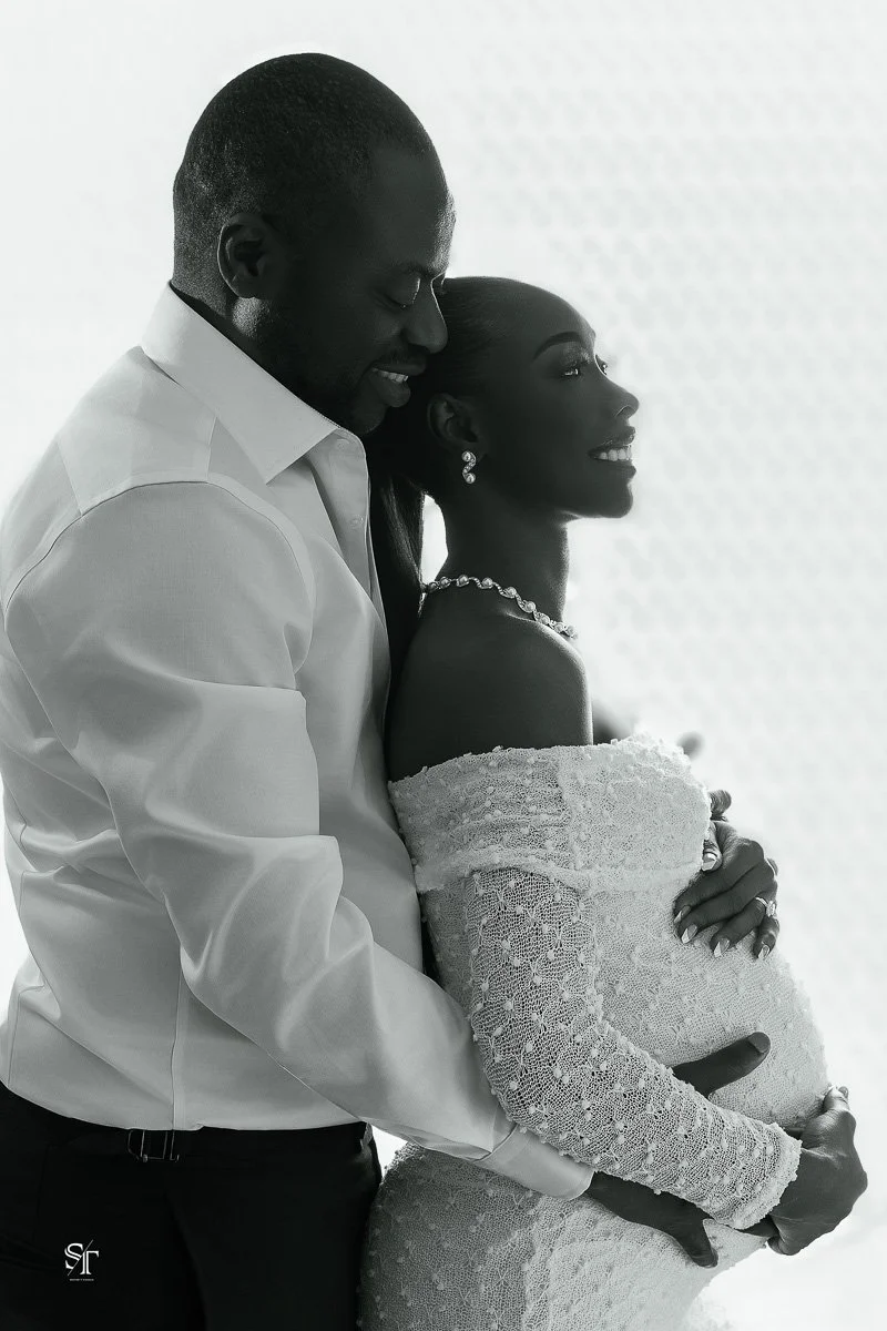 A couple dressed in wedding attire embracing, with the man wearing a white shirt and the woman in an off-shoulder lace dress and jewelry, in a black and white photograph.